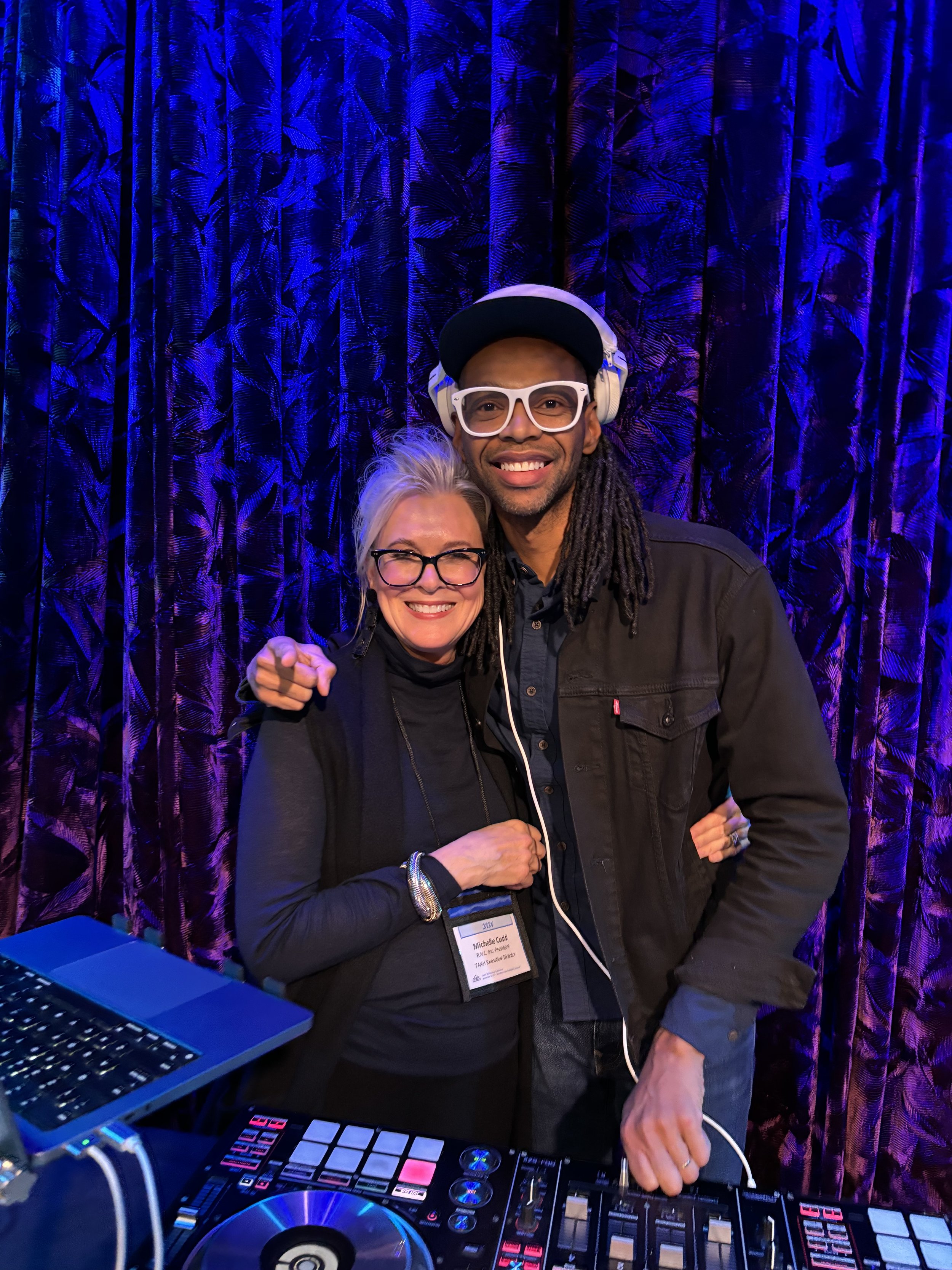Two people smiling, standing near DJ equipment. One is wearing headphones and a cap, the other has glasses. The background is a dark purple curtain.