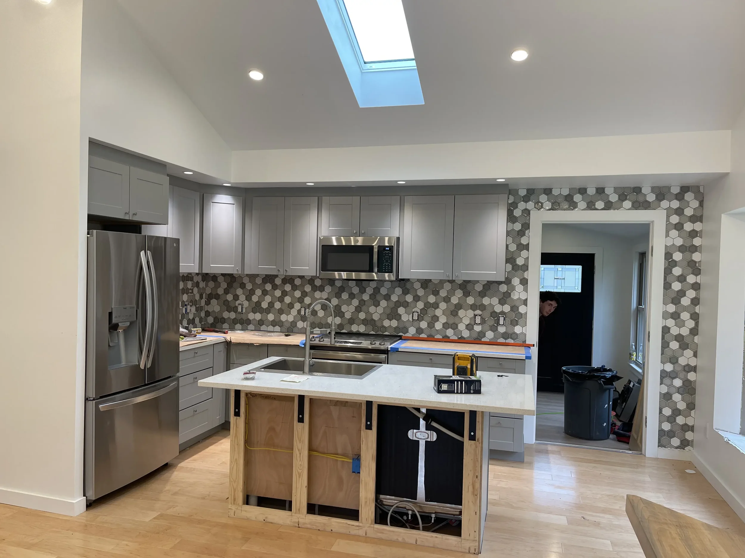 Kitchen under construction with gray cabinets, a stainless steel refrigerator, a microwave, a new island with a white countertop, and a gray hexagon tile backsplash. A man is partially visible in the doorway to the back room.