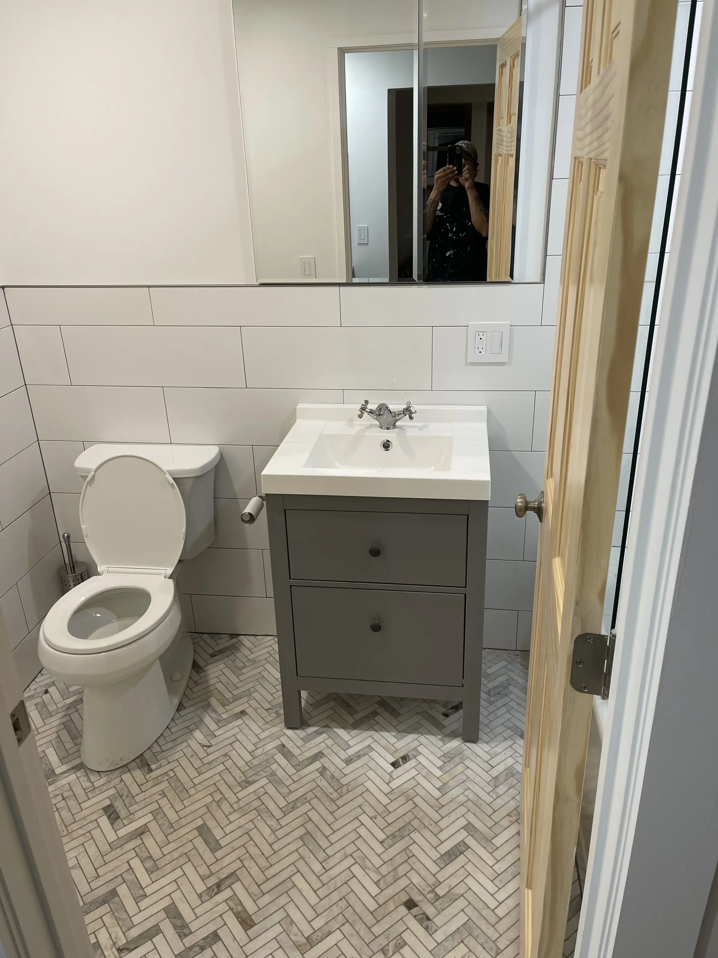 Small bathroom with white tiled walls, a toilet, a gray vanity with a white sink, and a large mirror above the sink. A person is visible in the mirror taking a photo. The floor has a herringbone tile pattern.