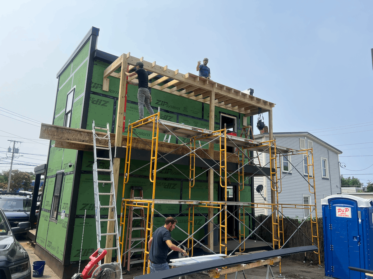 Construction workers building the second floor of a house with green exterior sheathing, scaffolding, and a ladder.