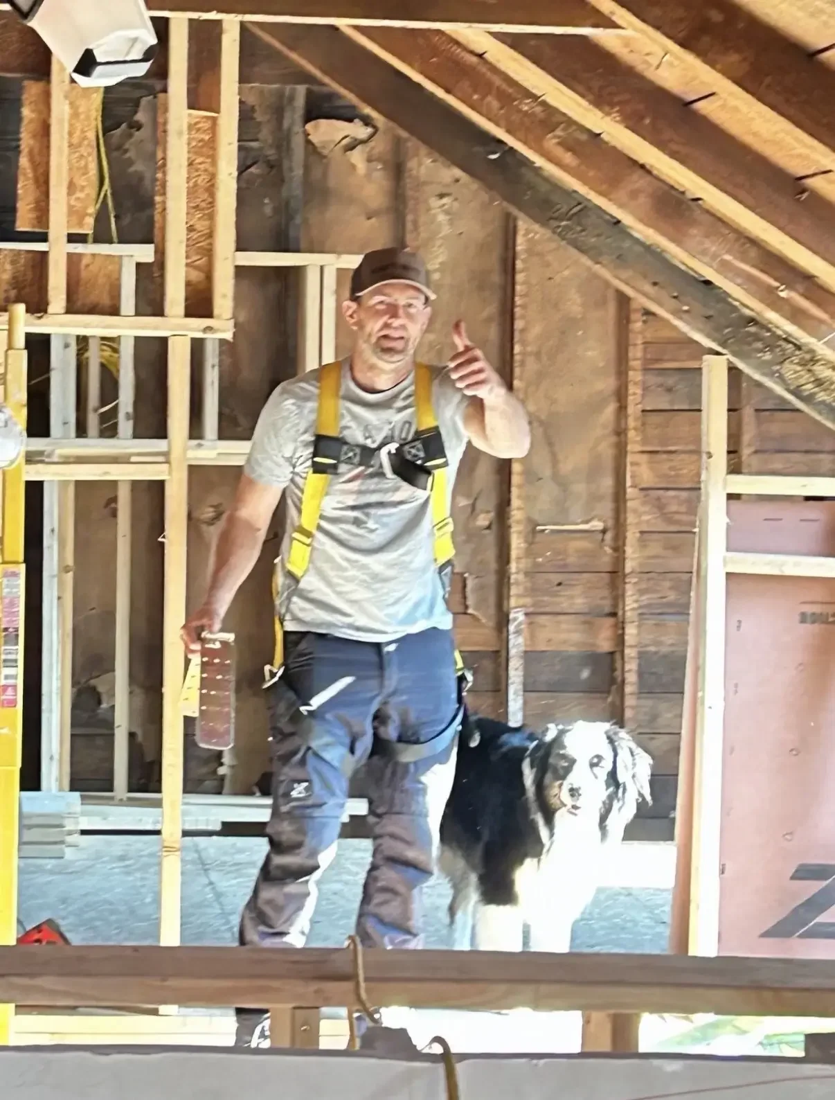 A man wearing a grey t-shirt, yellow harness, and camouflage pants standing inside a construction site with exposed wooden framing and roof trusses, pointing and smiling while holding a water bottle, beside a black and white dog.