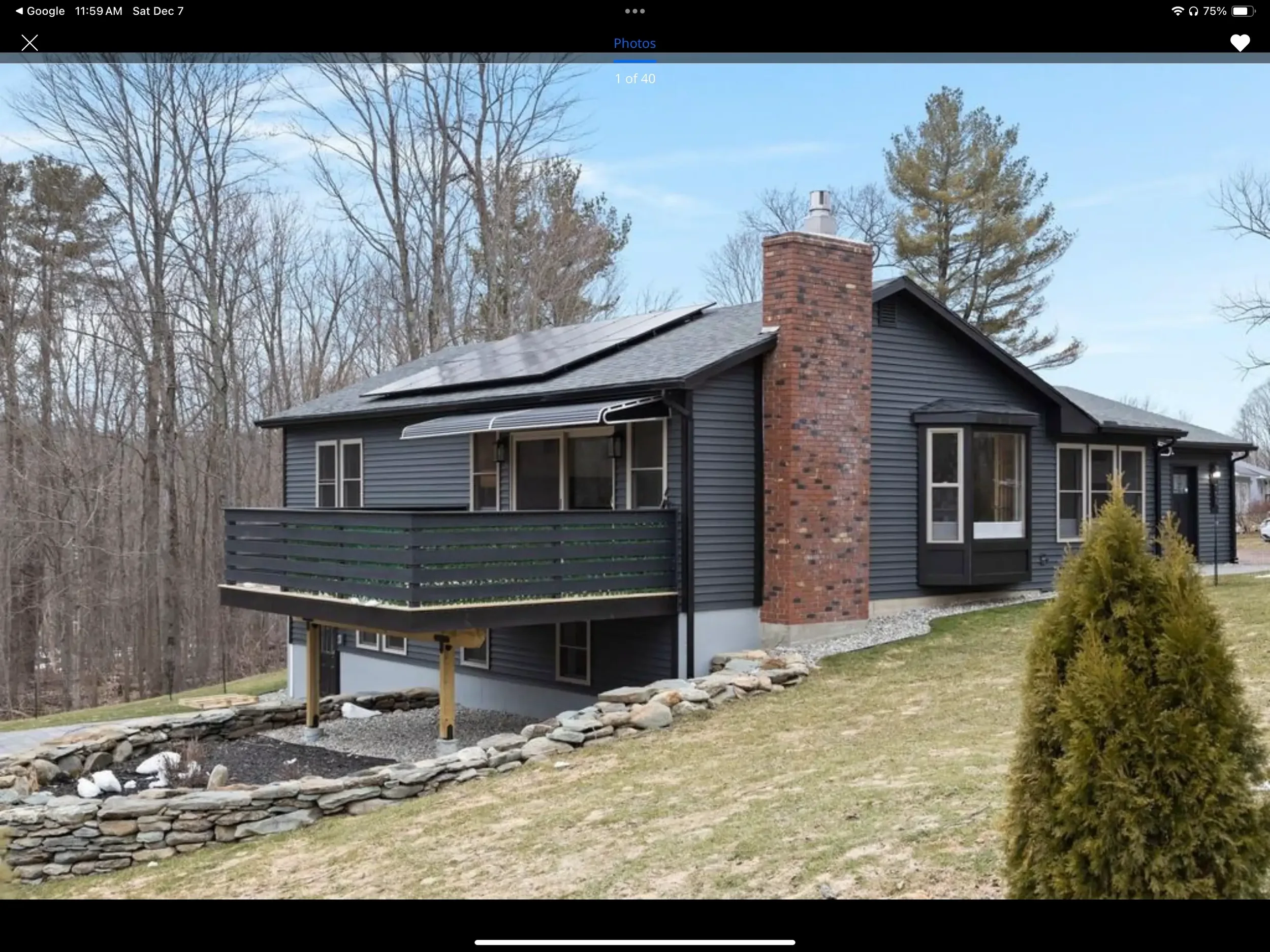 Black house with a brick chimney, solar panels on the roof, and a small deck with a railing overlooking a sloped lawn and trees.