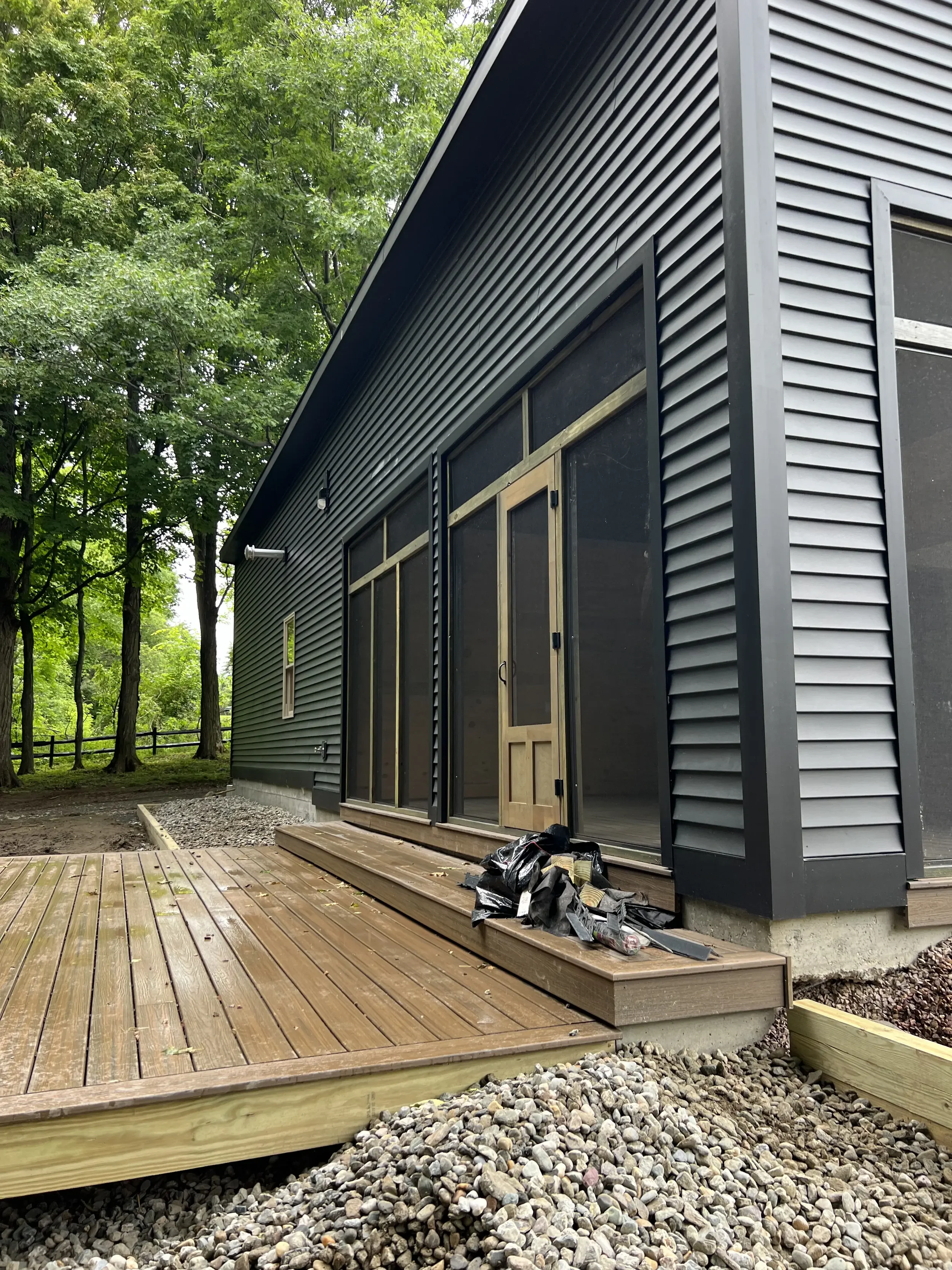 View of a newly constructed wooden deck attached to a modern black house with large sliding glass doors, surrounded by trees.