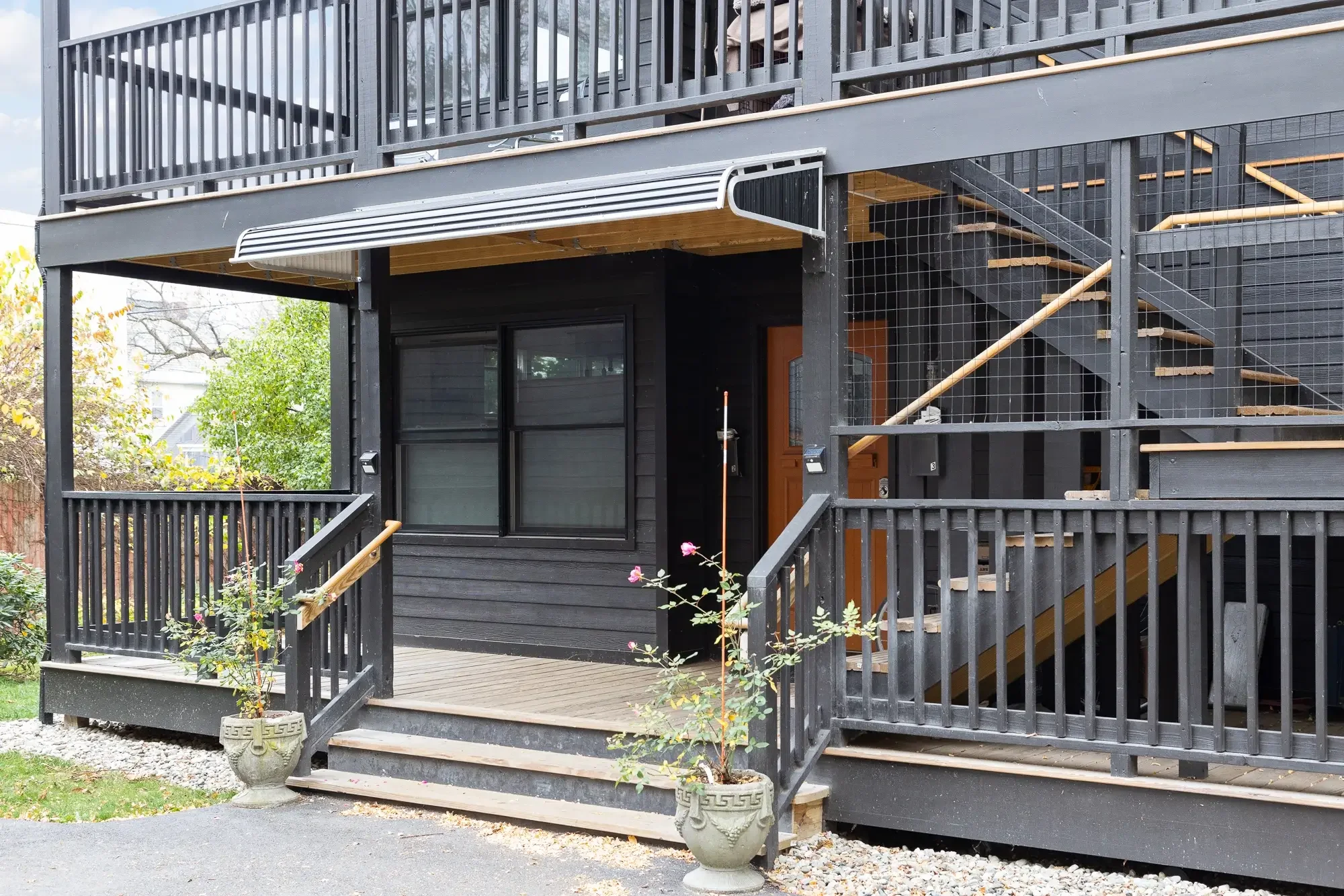 Exterior view of a two-story black wooden house with a deck, staircase, and railing, with potted plants and trees visible.
