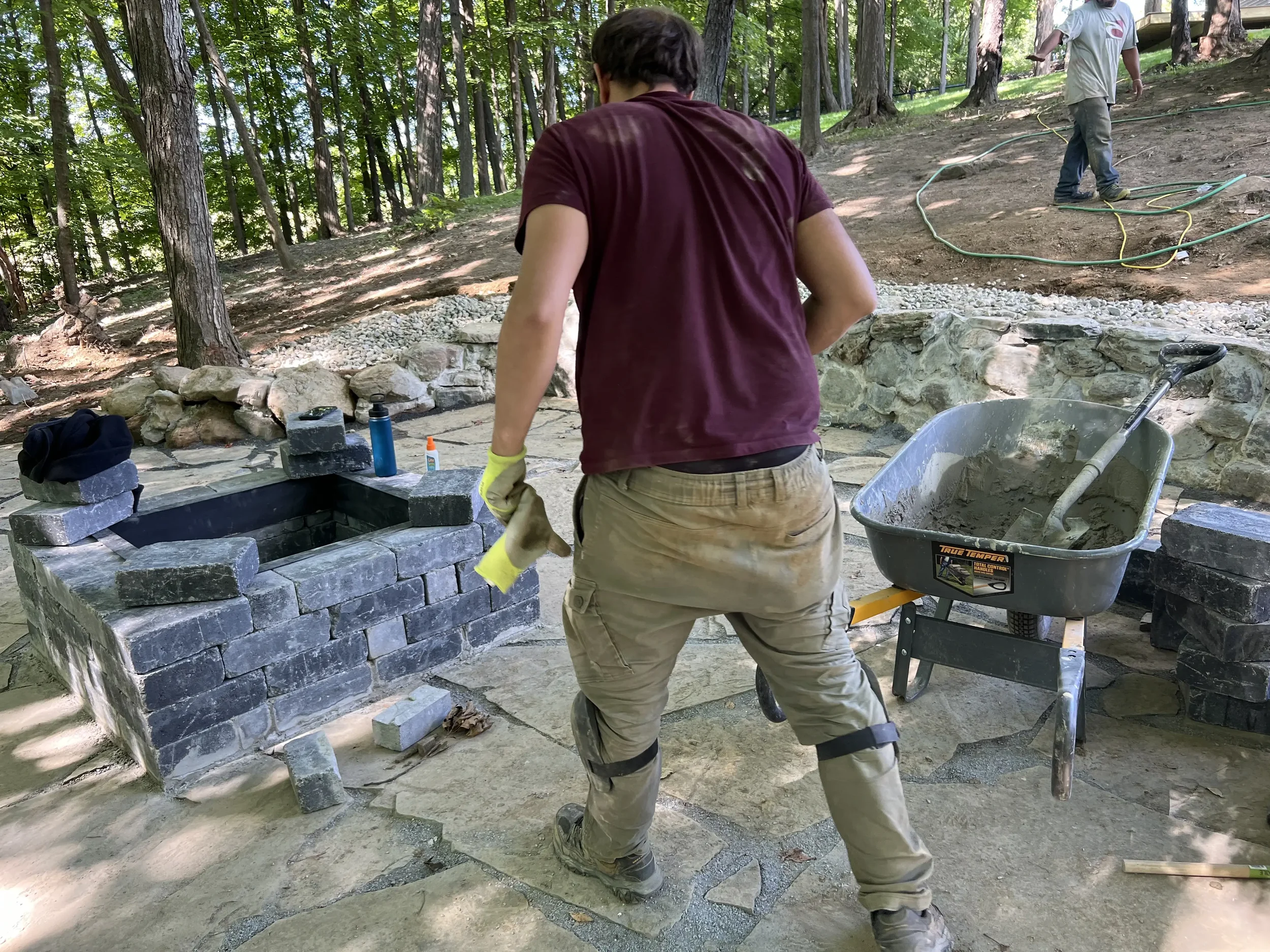 Person building a stone structure outdoors, with tools and a wheelbarrow nearby, in a wooded area.