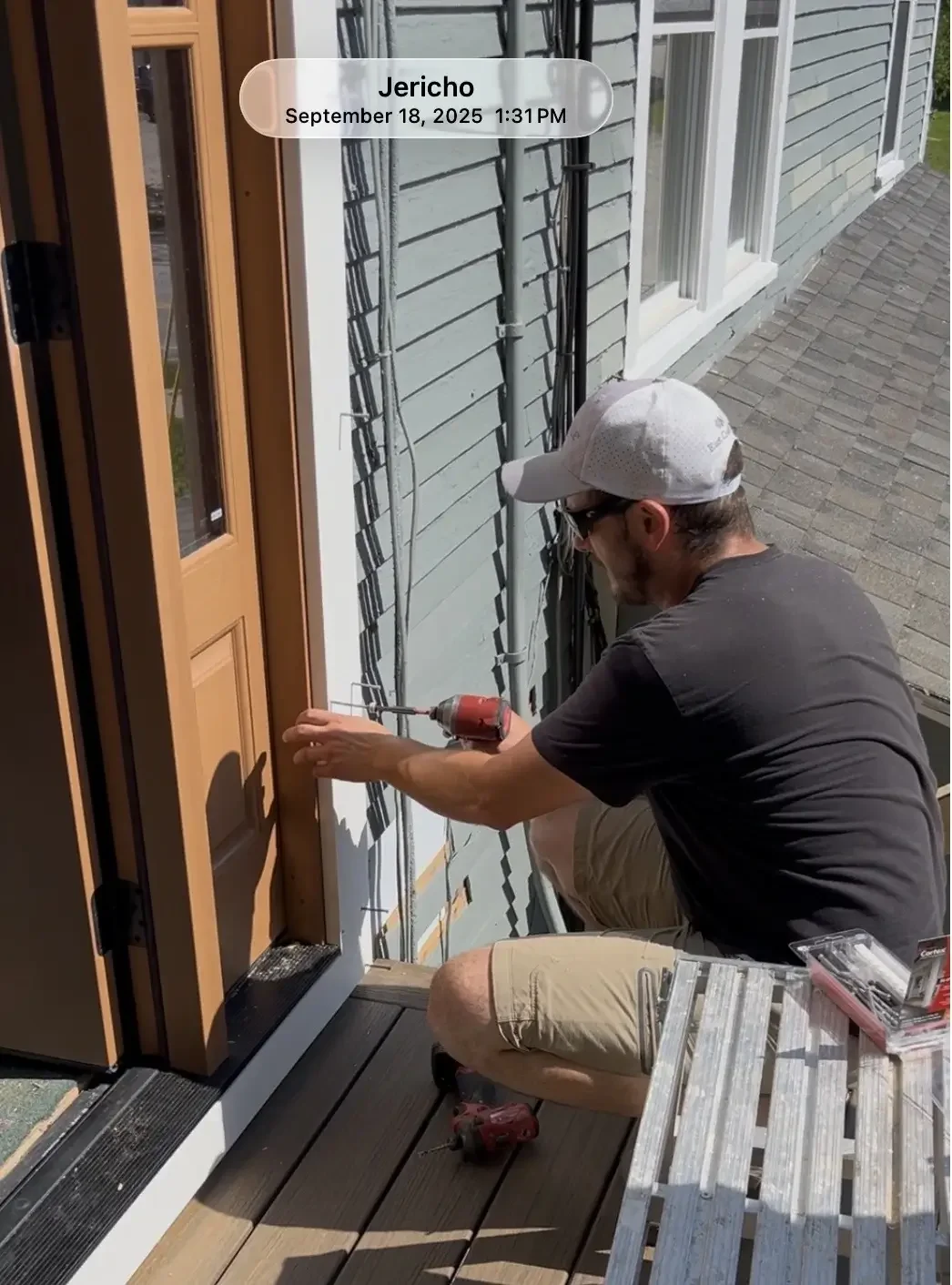 A man kneeling on a porch installing or repairing a doorframe, using a cordless screwdriver, with tools in a plastic container nearby.