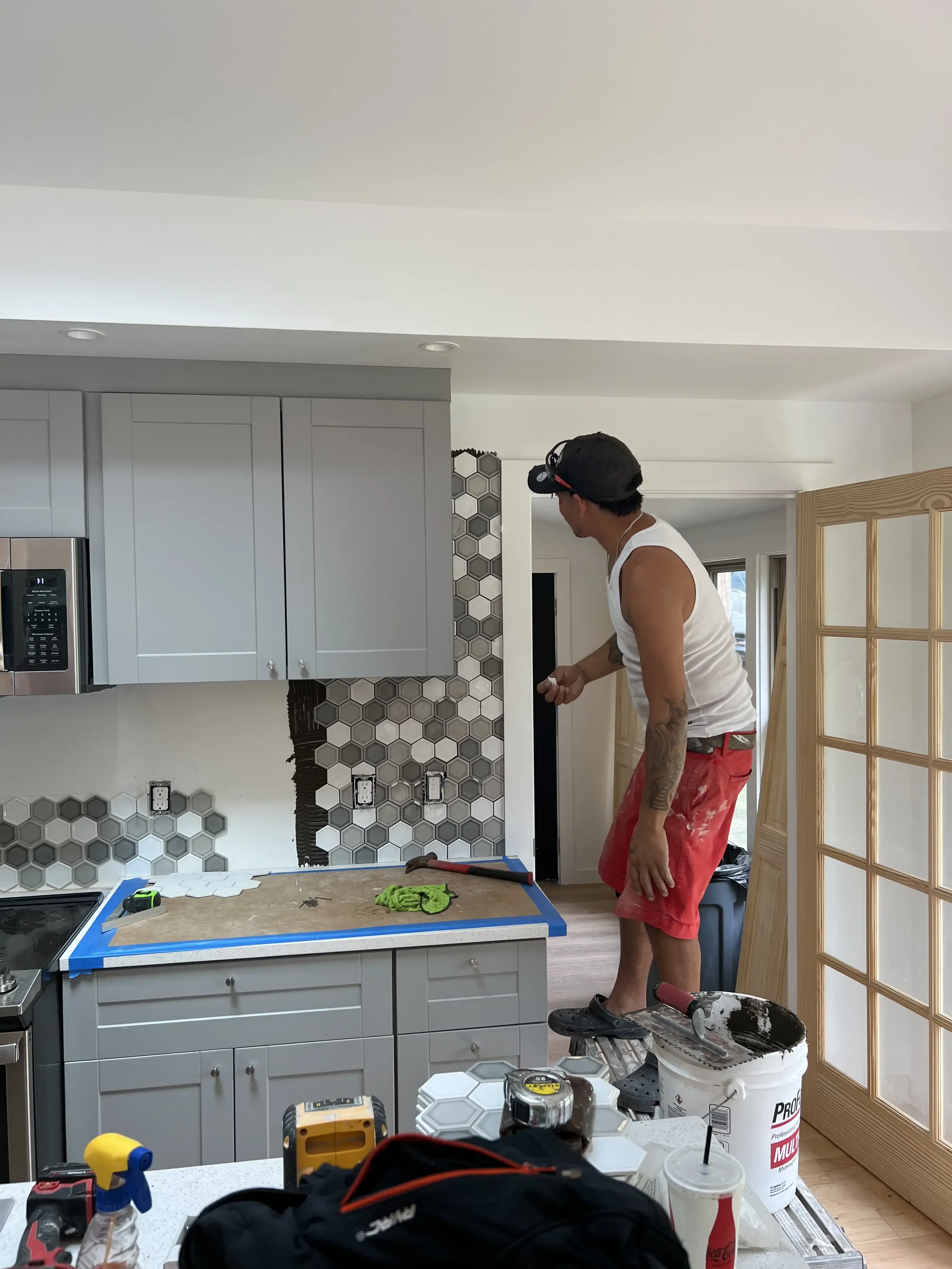 A man wearing a black cap, white tank top, and red shorts is working on installing or repairing tiles on a kitchen wall. The kitchen has gray cabinets, and there are various tools and materials scattered on the counter and floor.