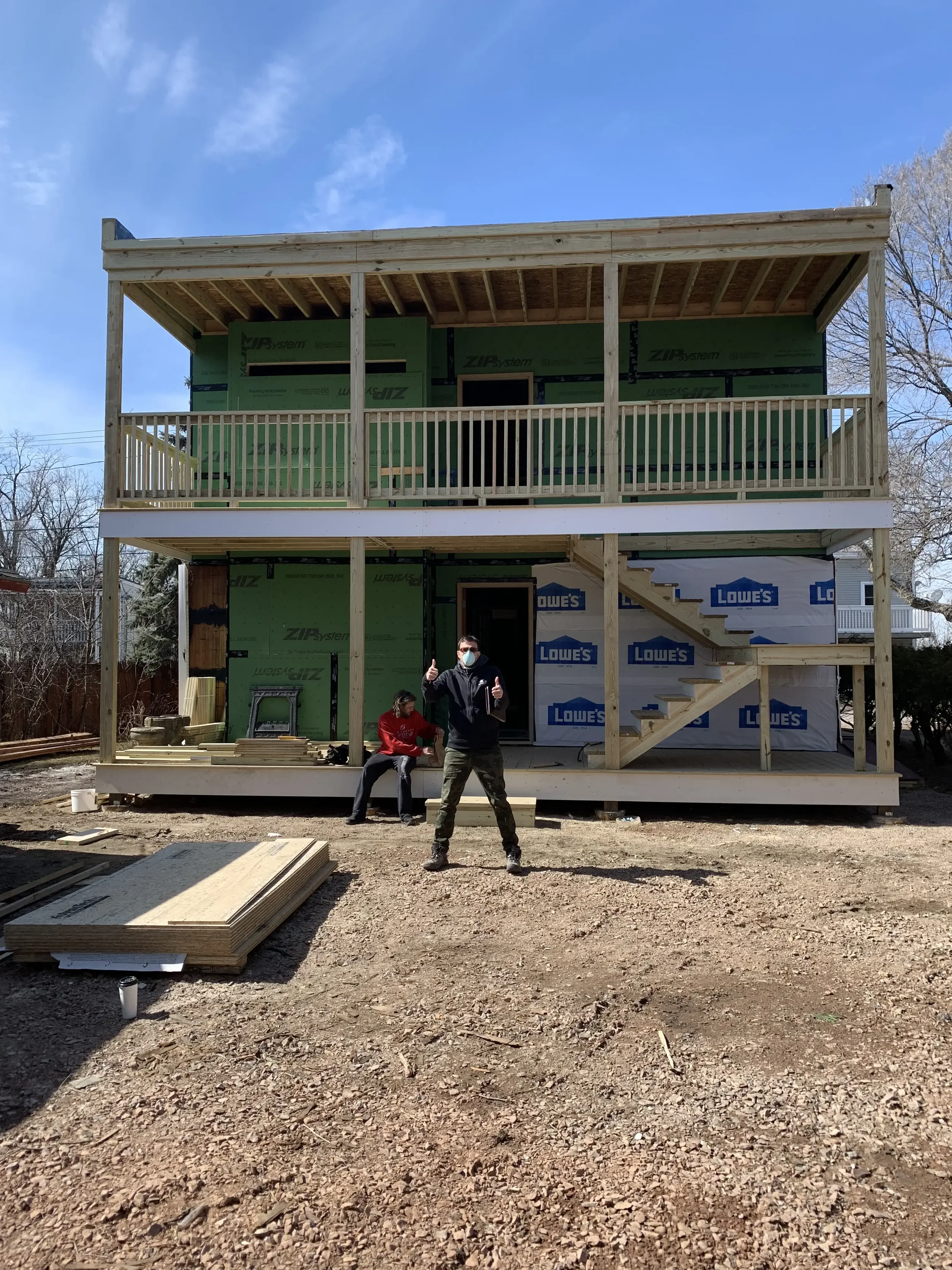 Two people in construction gear working on building a two-story house with an open front and stairs, set outdoors on a dirt lot.