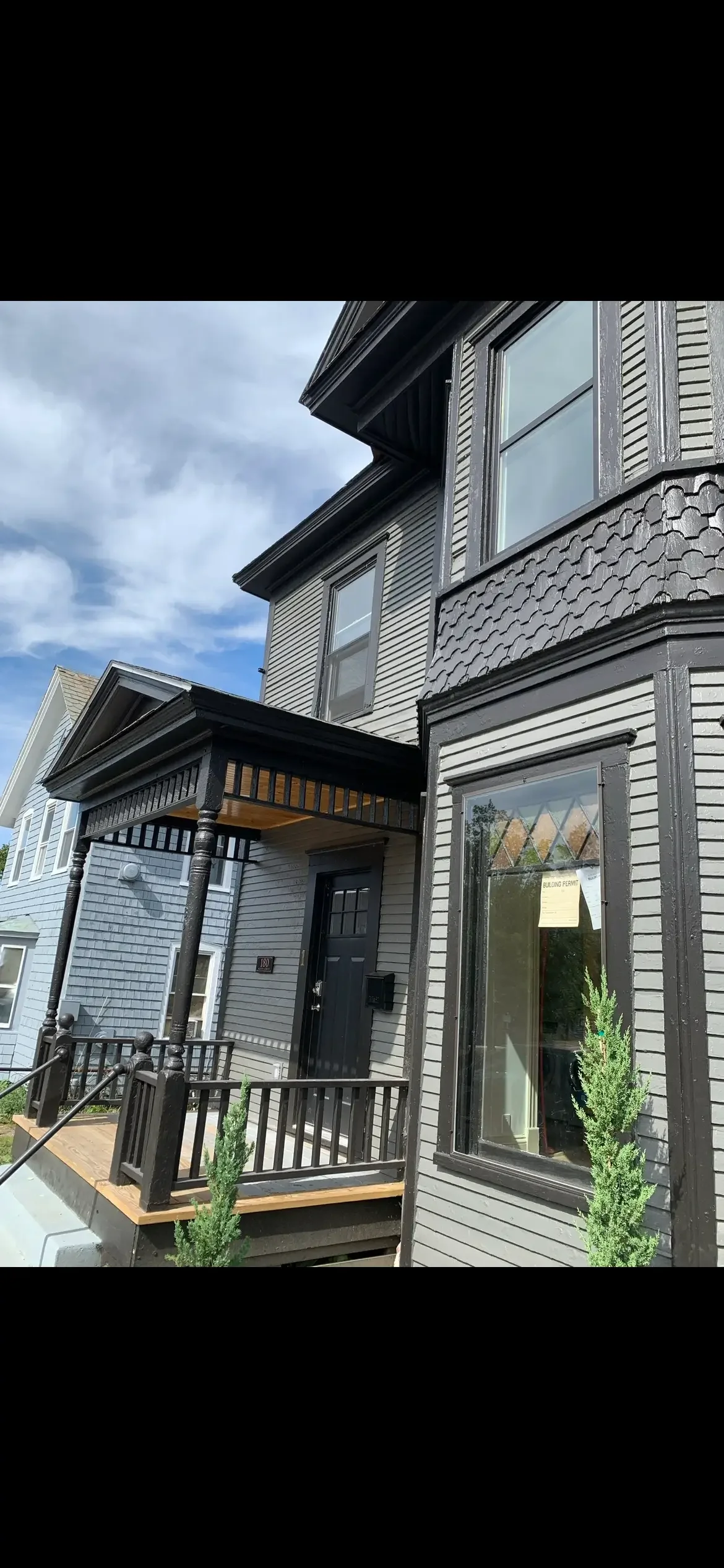 Exterior view of a two-story house with black trim, black door, front porch, metal railings, and small green shrubs near the porch, under a partly cloudy sky.