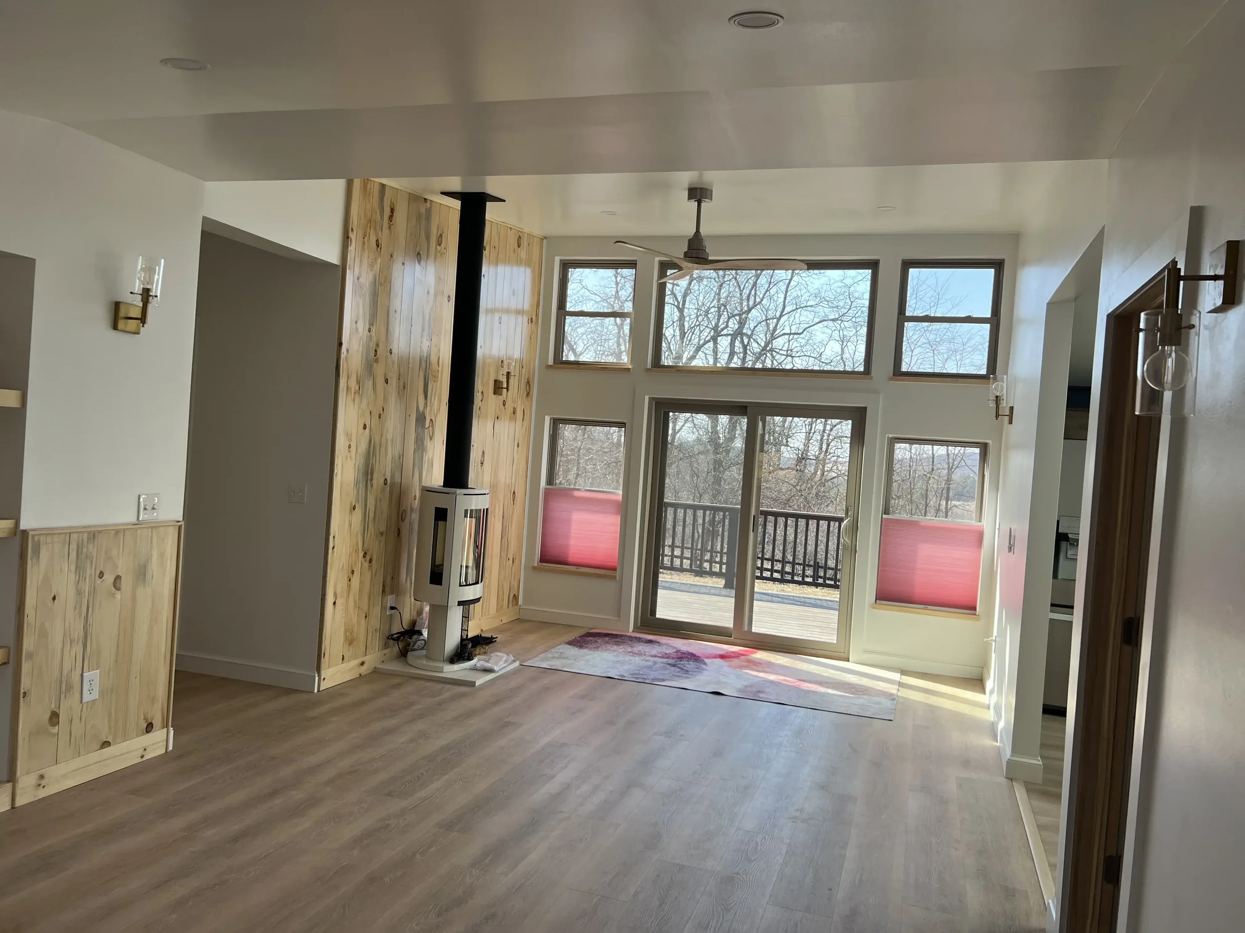 Empty living room with large windows, a wood stove, wooden accents on the wall, and a sliding glass door leading to a deck.