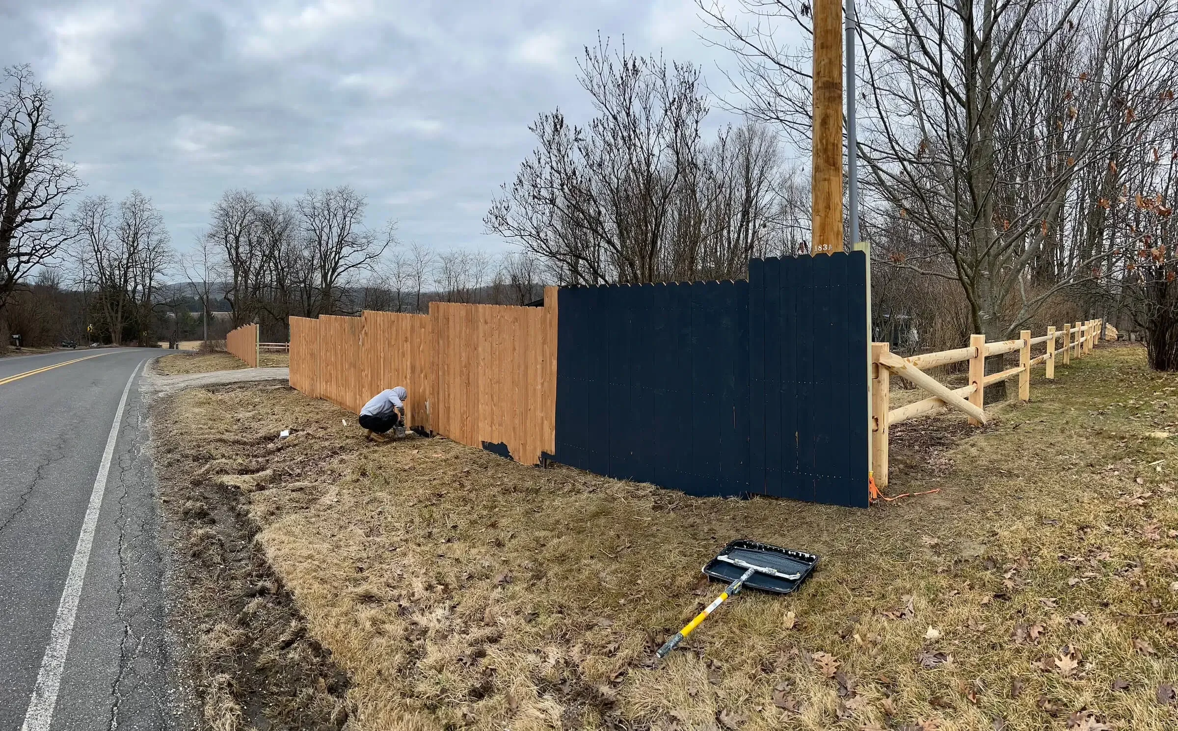 Person installing a wooden privacy fence along the roadside on a cloudy day, with a wheel roller lying on the grass.