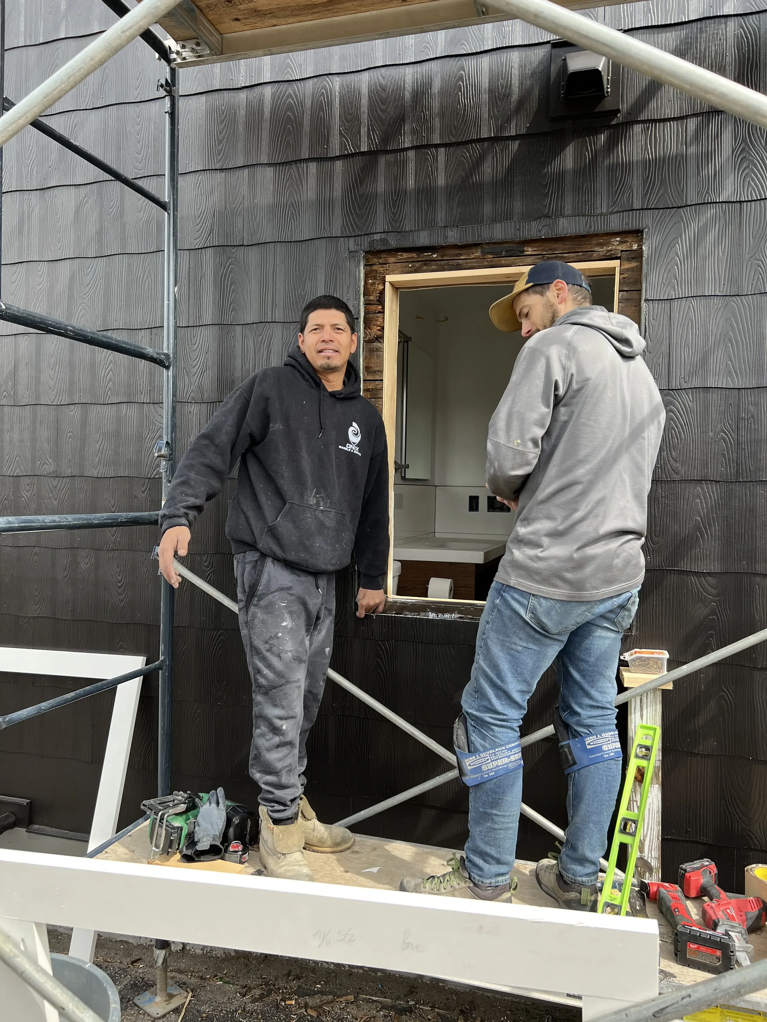 Two construction workers standing on a scaffold in front of a house wall, with one worker smiling and the other looking down, surrounded by tools.