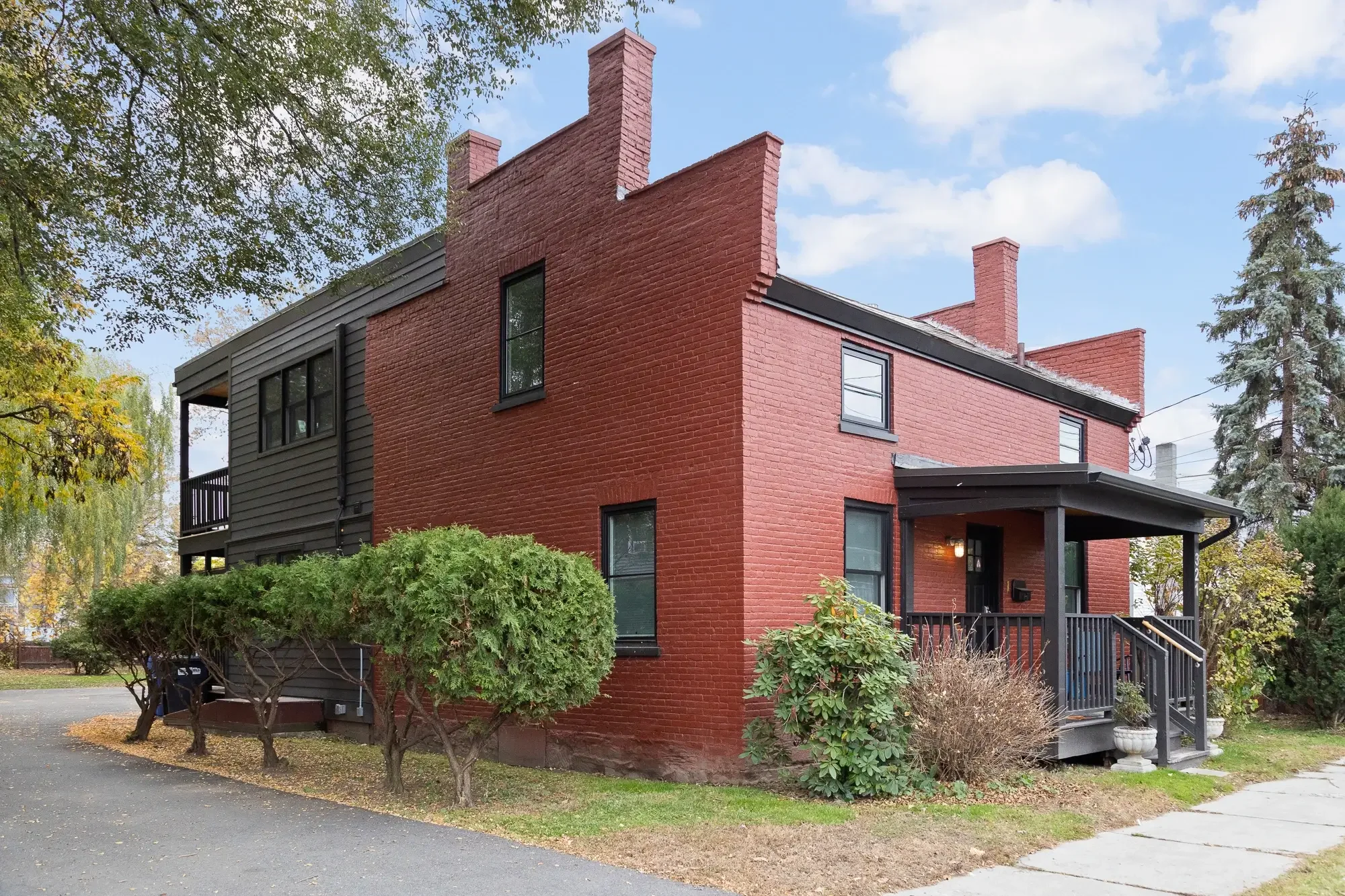 A two-story brick house with black window frames and a black porch, surrounded by shrubbery and trees, under a partly cloudy sky.