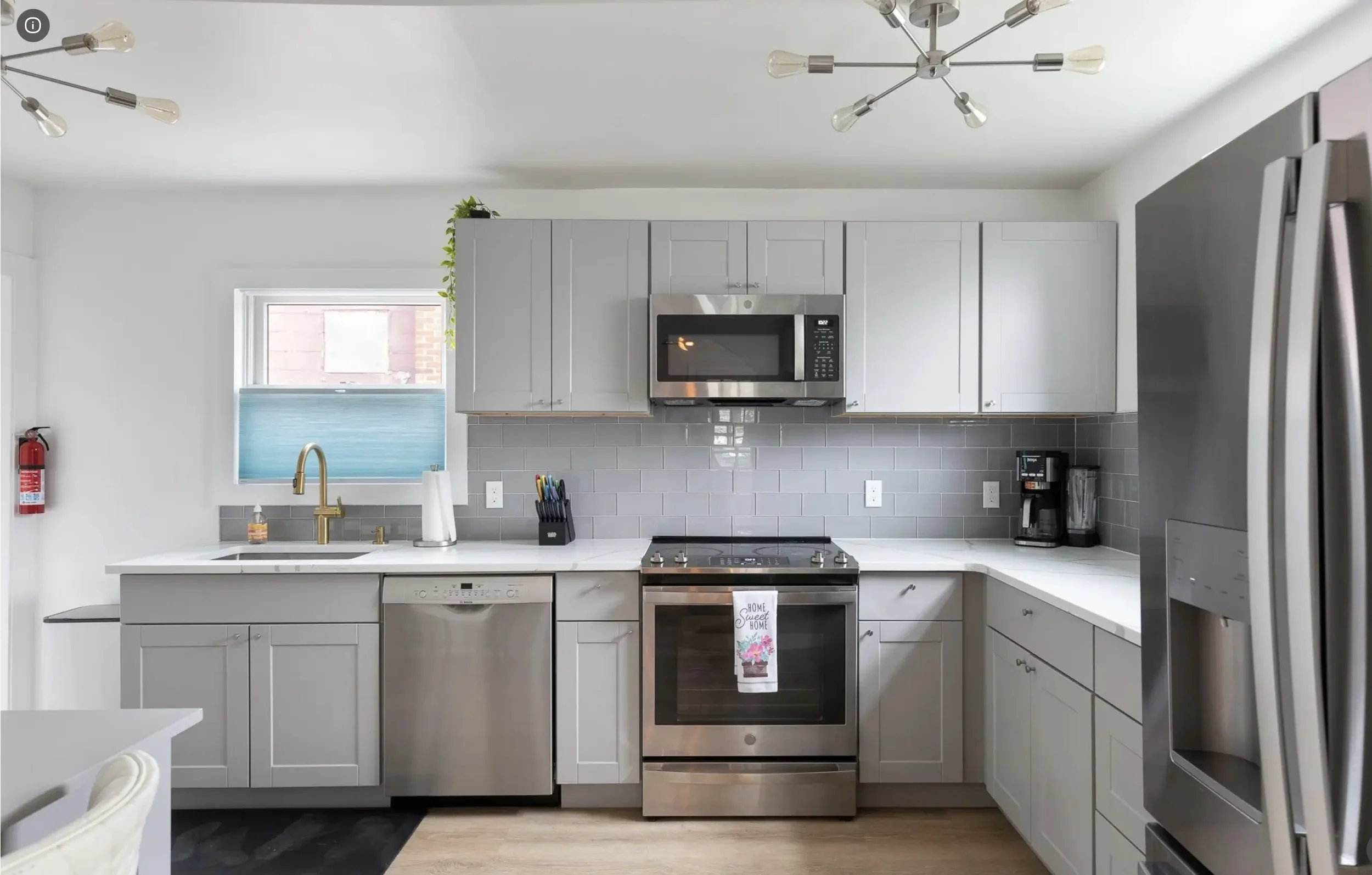 Modern kitchen with gray cabinets, stainless steel appliances, including microwave, oven, dishwasher, and refrigerator, a black coffee maker, a black utensil holder, and a window above the sink with a gold faucet.