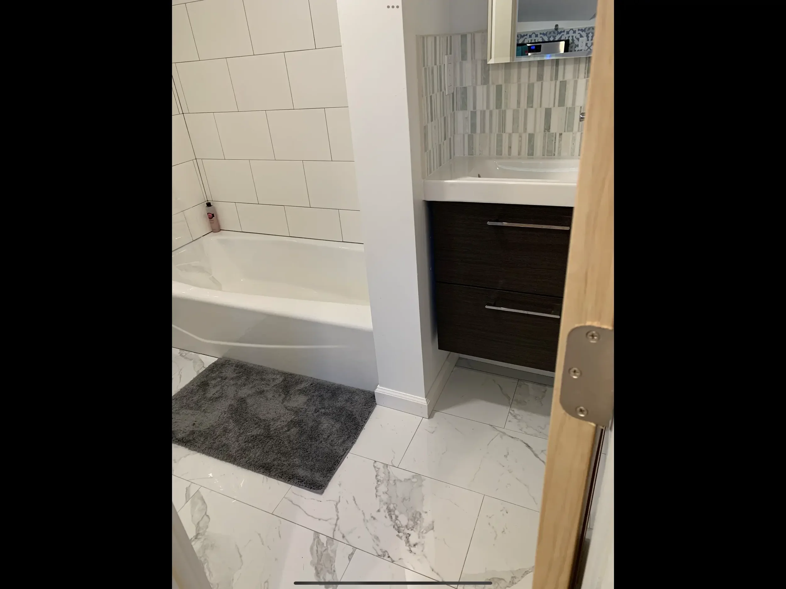 A bathroom with a bathtub on the left, a gray bath mat on the floor, and a vanity with a sink and dark cabinet on the right. The wall behind the vanity has light-colored tiles with a striped pattern.