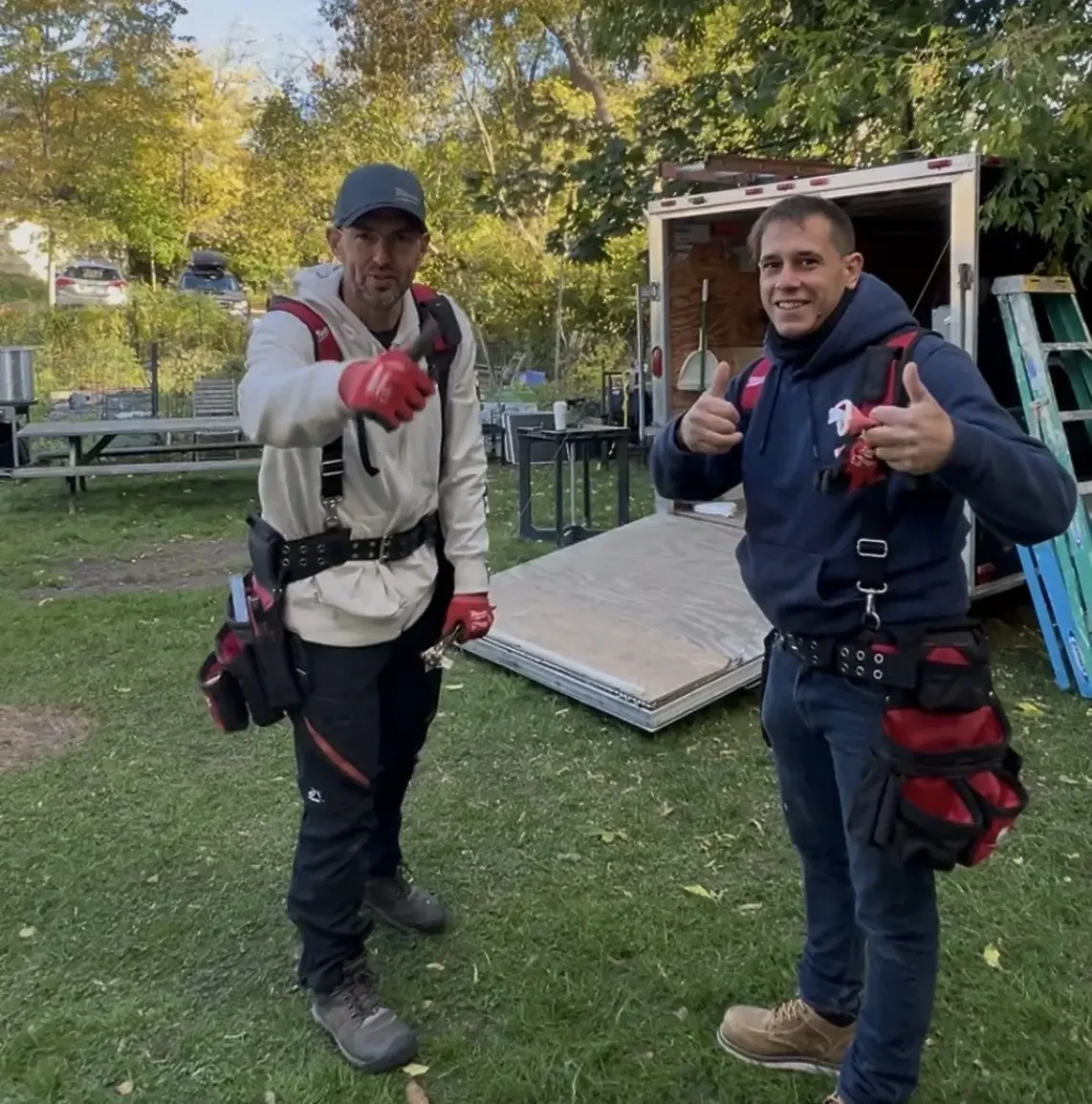 Two men standing outdoors in front of a work trailer with tools and materials inside. One man is wearing a white jacket and red gloves, and the other is wearing a blue jacket and tan boots. Both are smiling and giving thumbs-up gestures.