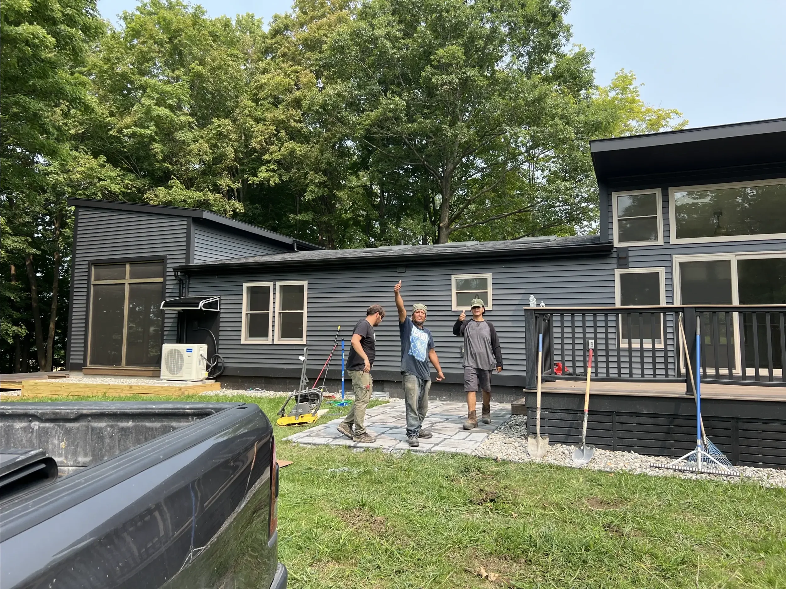 Three men working outside on a residential construction project, installing a stone walkway in the backyard of a modern house with black siding and multiple large windows, surrounded by trees.