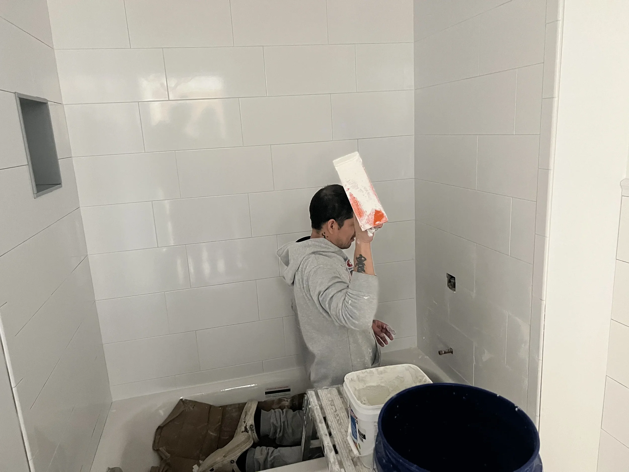 A man working on tiling a bathroom wall, holding a trowel, with buckets of grout nearby.