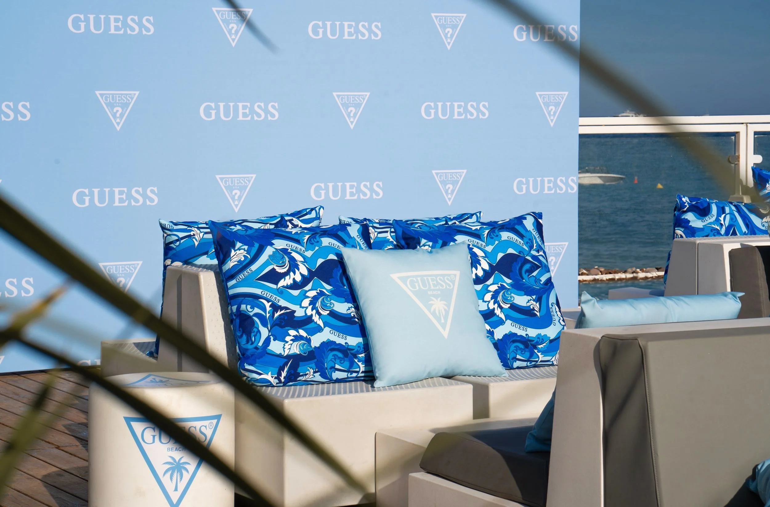 Coussins bleus avec logo Guess sur des sièges extérieurs près de la plage.