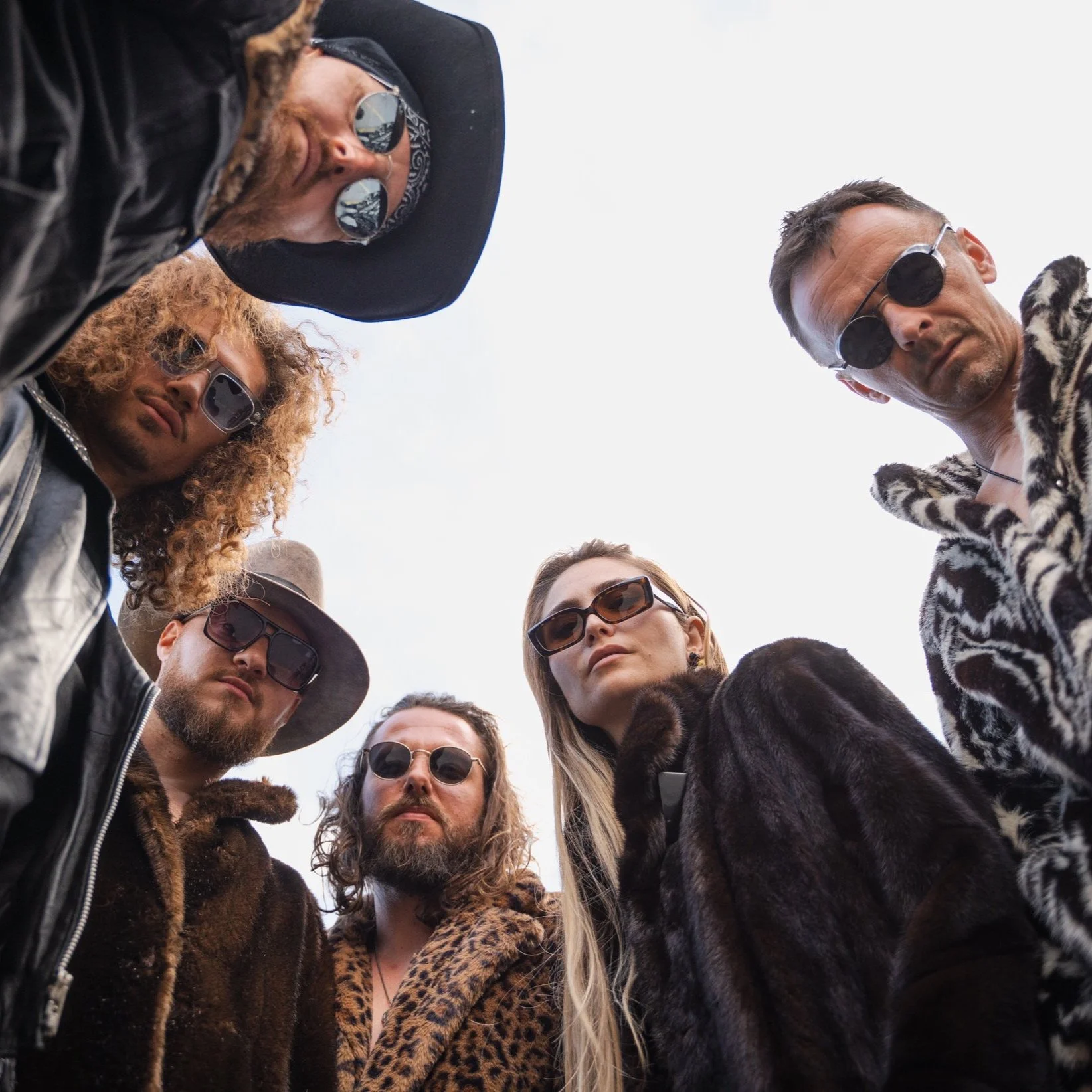 Un groupe de six personnes portant des lunettes de soleil et des manteaux, regardant vers le bas. Style rétro et décontracté, ciel clair en arrière-plan.