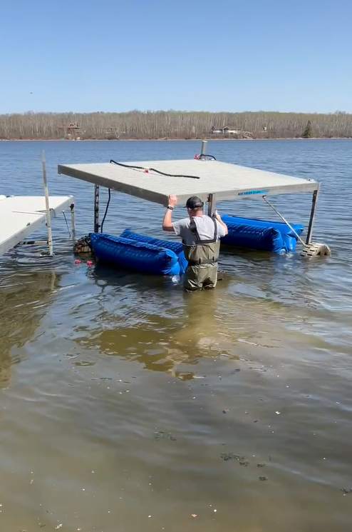 Person standing in water next to a floating dock with blue flotation devices, adjusting the structure, against a background of a lake and forest.