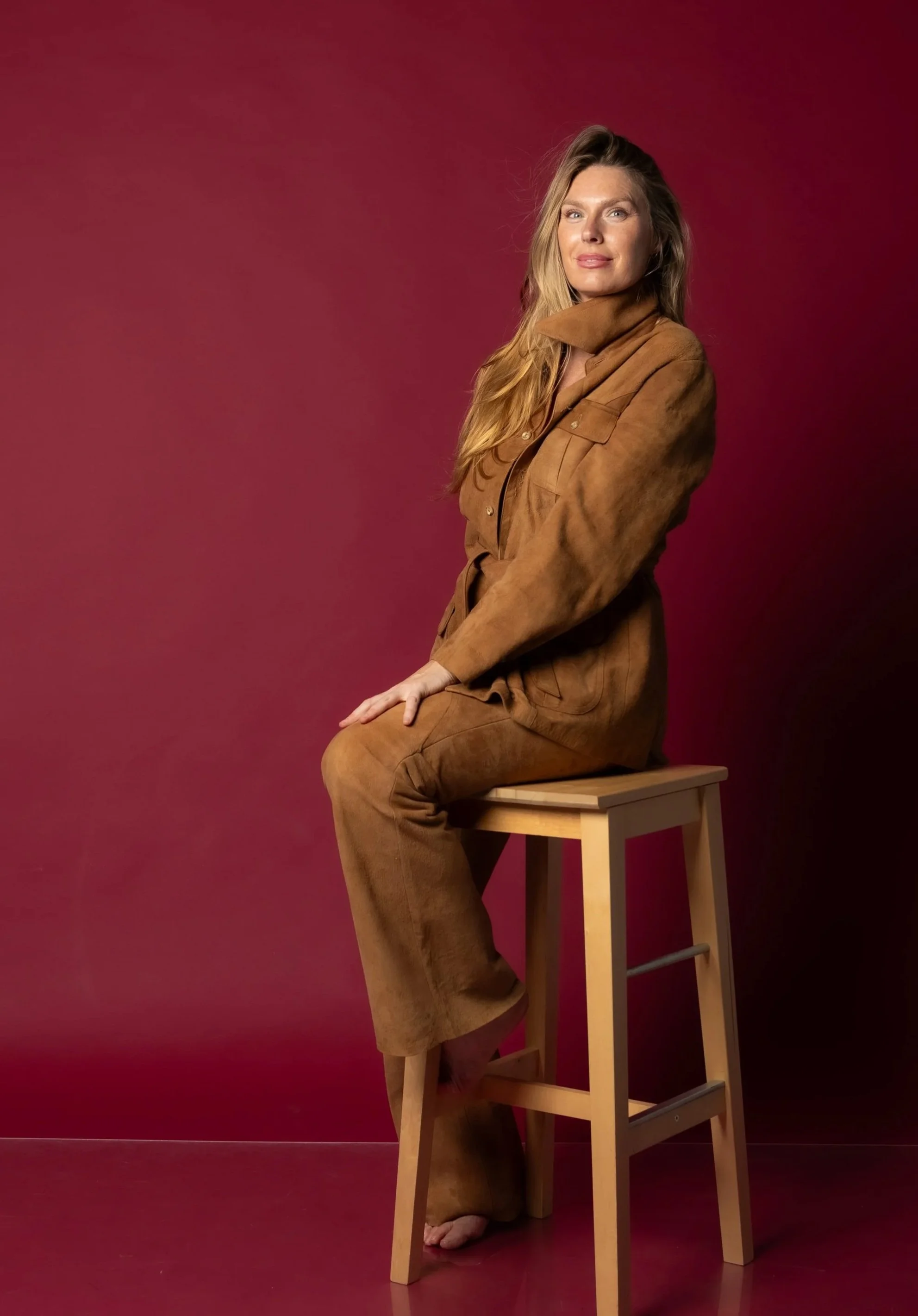 Ashlee Jowett - Contact me. A woman sitting on a wooden stool against a maroon background, wearing a brown suede outfit with long, wavy blonde hair.