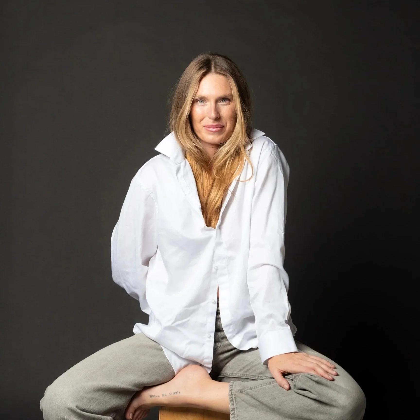 Ashlee Jowett - Contact me. A woman with long blonde hair wearing a white button-up shirt and beige pants, sitting with one leg crossed over the other against a dark background.