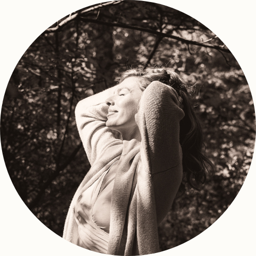 A sepia-toned image of a woman with closed eyes, arms raised behind her head, standing outdoors amidst trees.