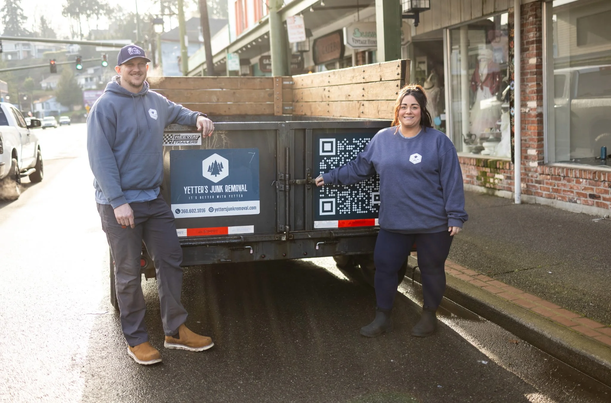 Two people stand beside a black trailer with a wooden load, parked on a city street. Both are smiling, with one man wearing a navy hoodie, gray pants, brown boots, and a navy cap, and a woman in a navy sweatshirt, dark pants, and boots. The trailer has a sign for Yetter's Junk Removal and a QR code.