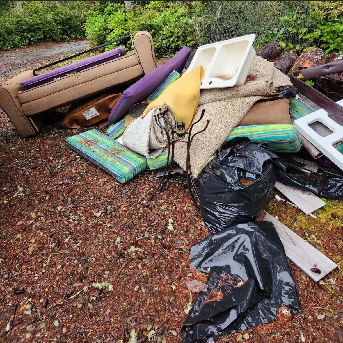A pile of discarded furniture and trash outdoors, including a broken sofa, mattress, plastic bags, a couch cushion, and various other debris on the ground surrounded by greenery.