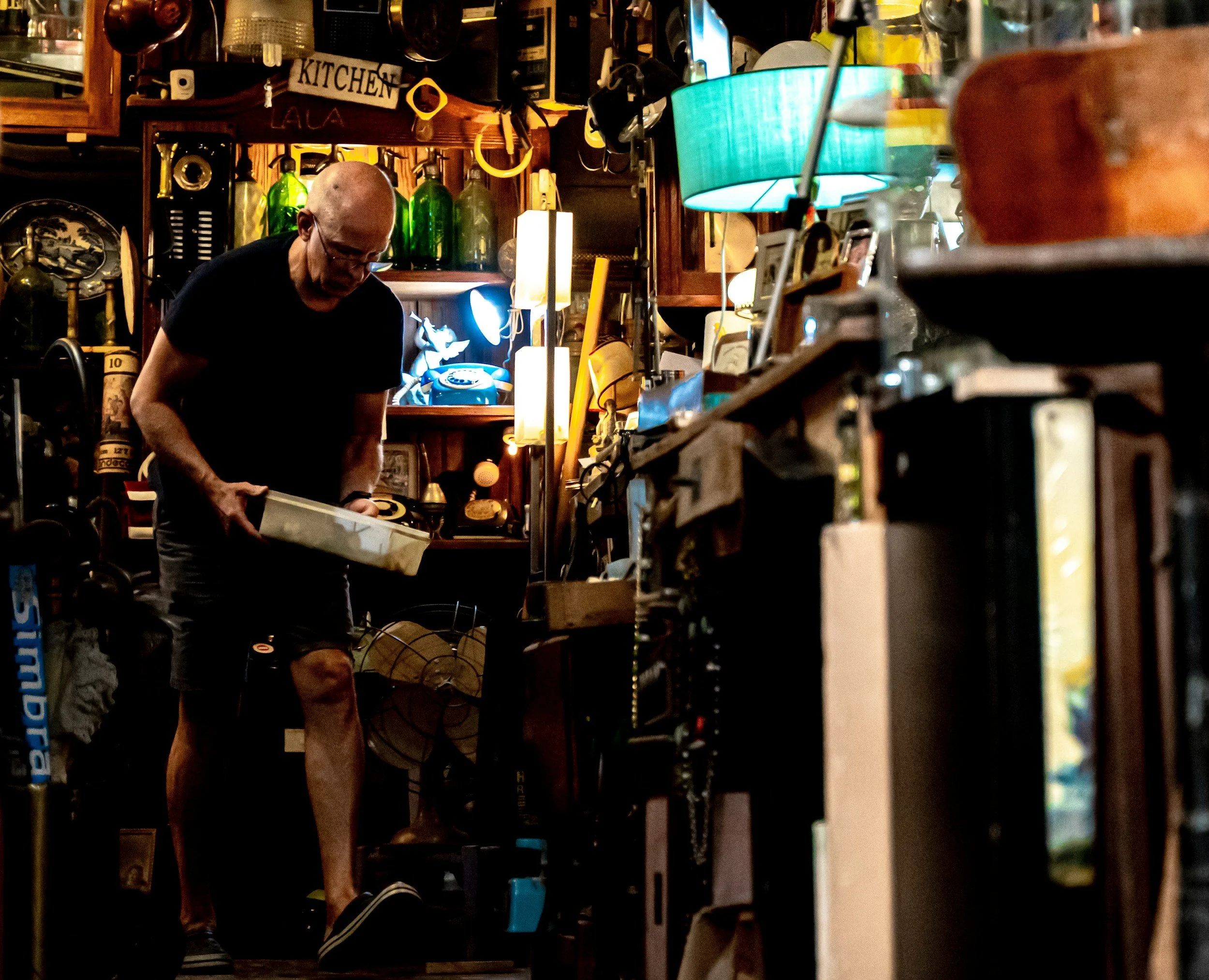 A person with a shaved head and glasses working in a cluttered workshop filled with various items such as lamps, bottles, and kitchenware.