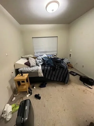Messy bedroom with an unmade bed, scattered clothes, and personal items on the floor and furniture.
