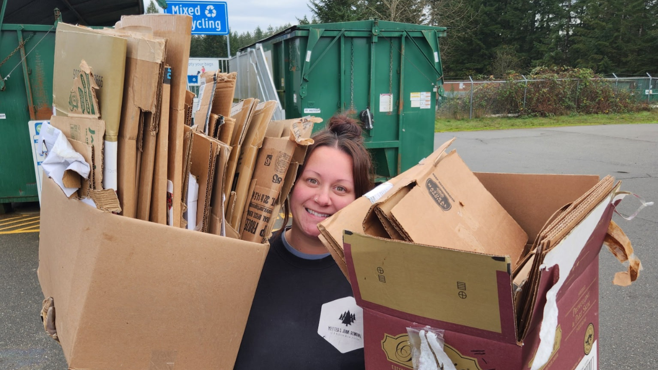 A smiling woman holding two large boxes filled with flattened cardboard boxes at a recycling center.