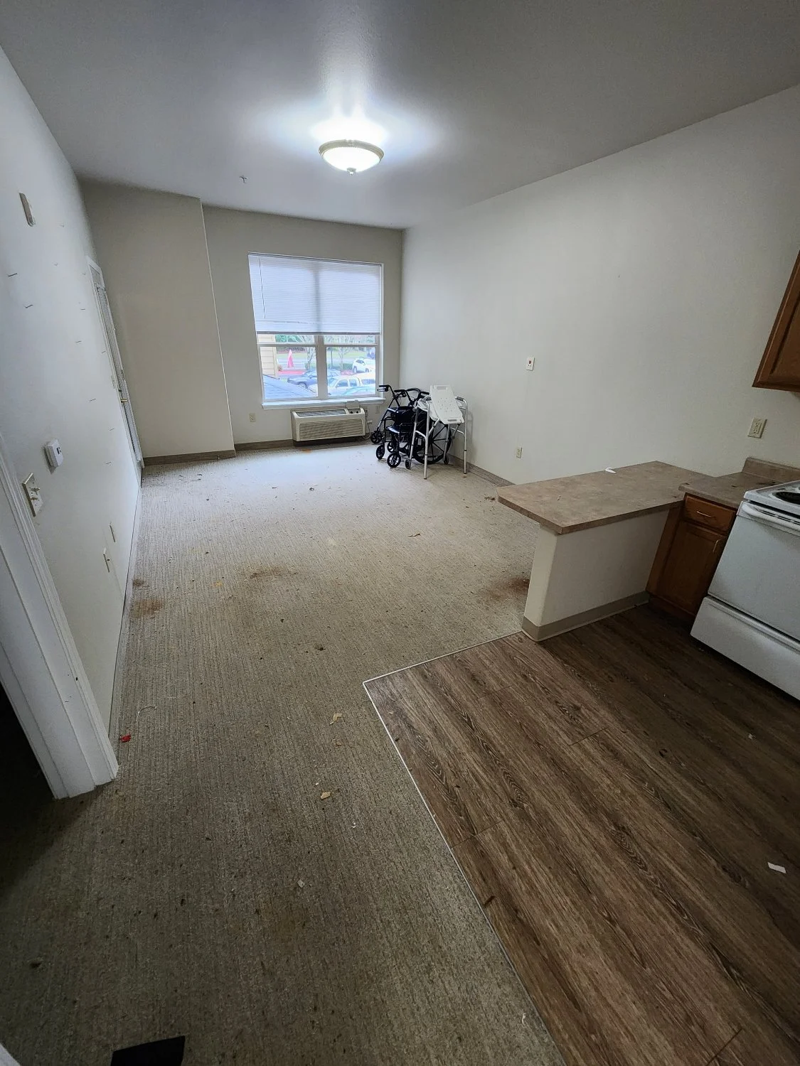 Empty apartment living room with dirty carpet, some small debris, a window with closed blinds, a few wheelchairs, and an open kitchen area with a countertop.