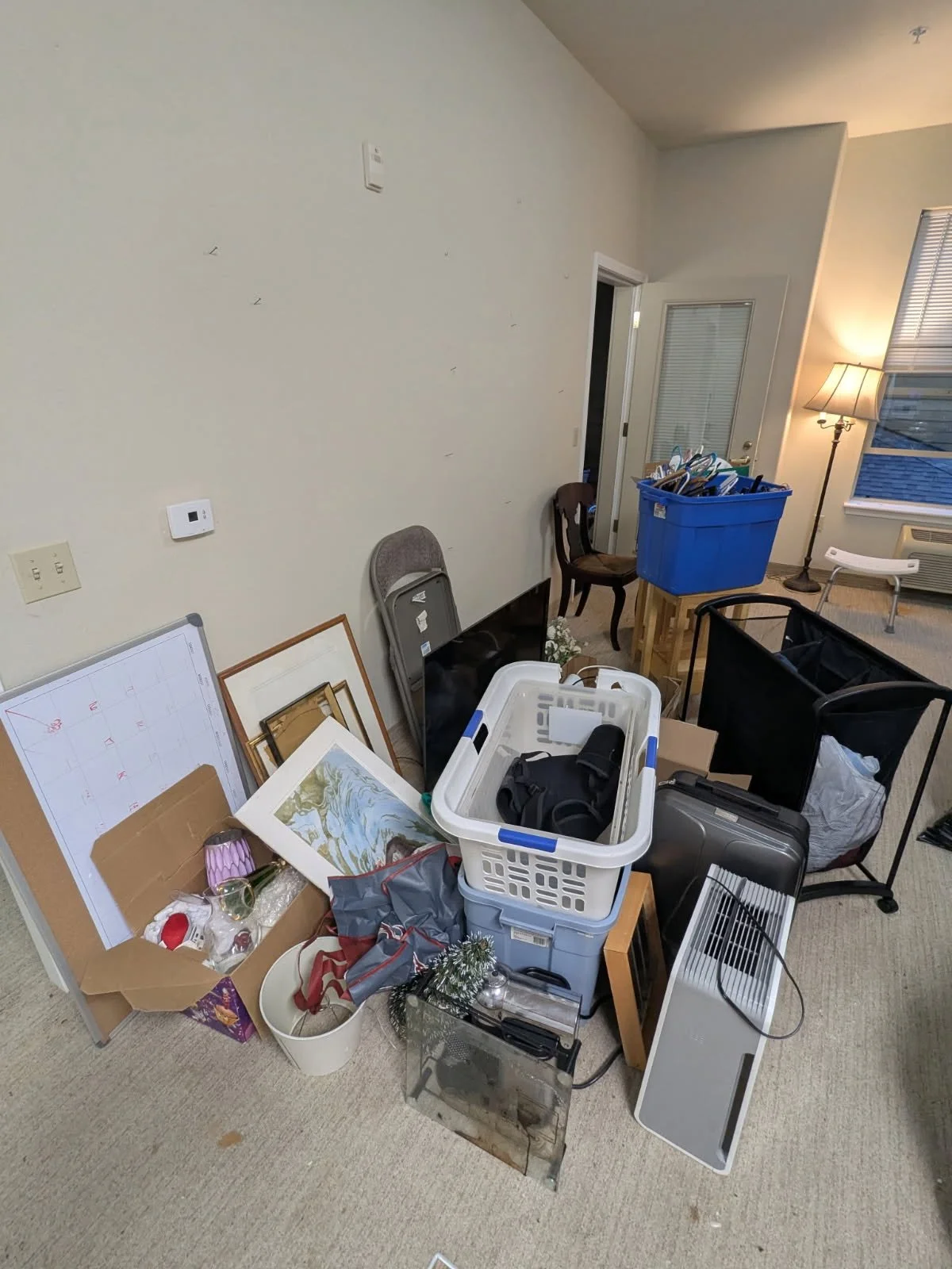 Cluttered room with various household items, furniture, boxes, and decorations, including a picture frame, a blue storage bin, a lamp, and a fan.