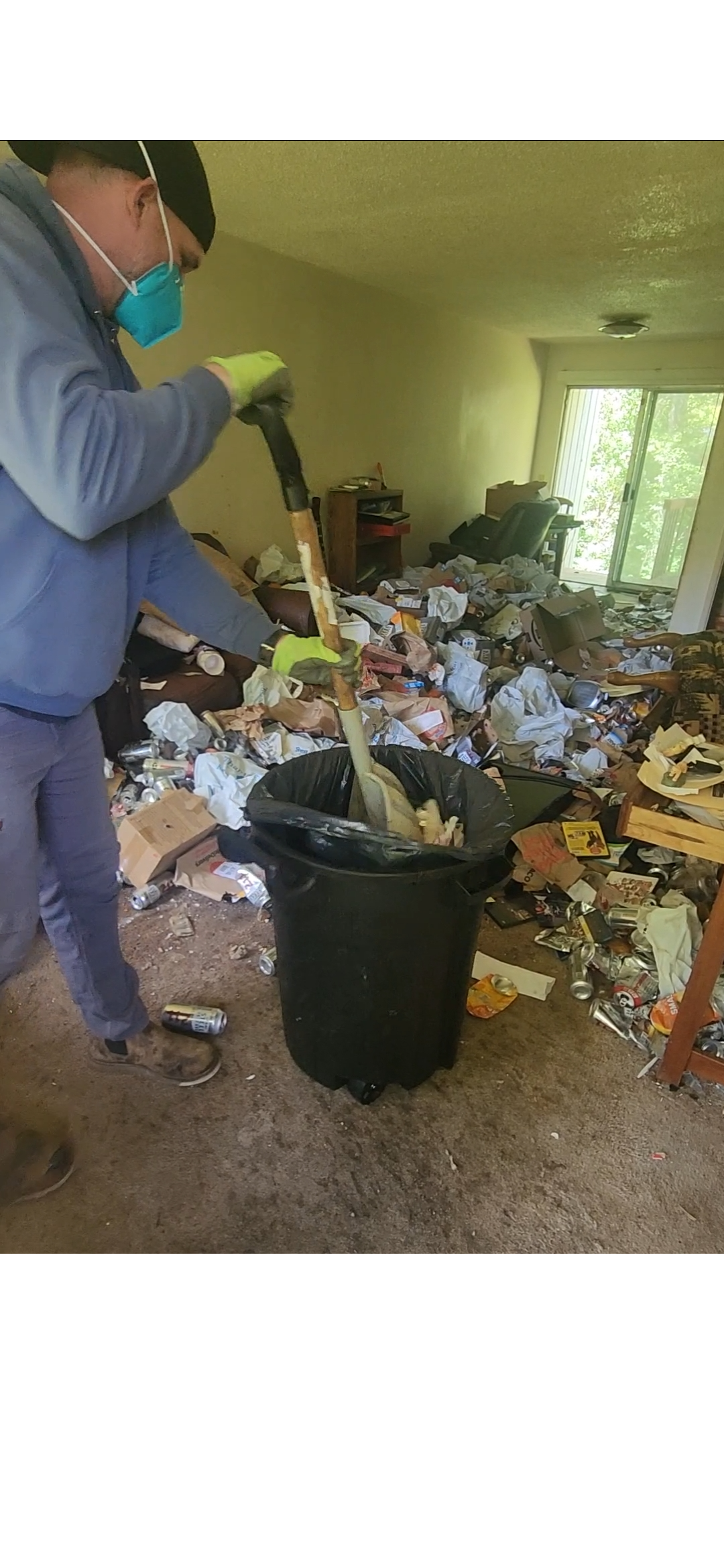 A person wearing a face mask, gloves, and a beanie is cleaning clutter in a living room, throwing trash into a trash can amid scattered trash, paper, and cans.