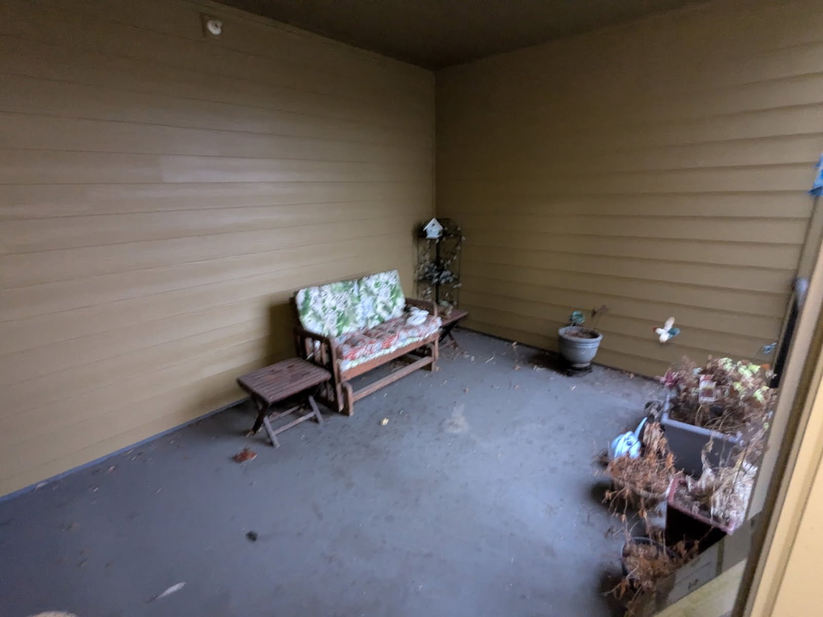 A small outdoor porch area with wood paneled walls and a concrete floor. There is a wooden bench with floral cushions, a small wooden side table, a metal shelving unit, a large flowerpot with no plant, and a window box with dried plants, along with s
