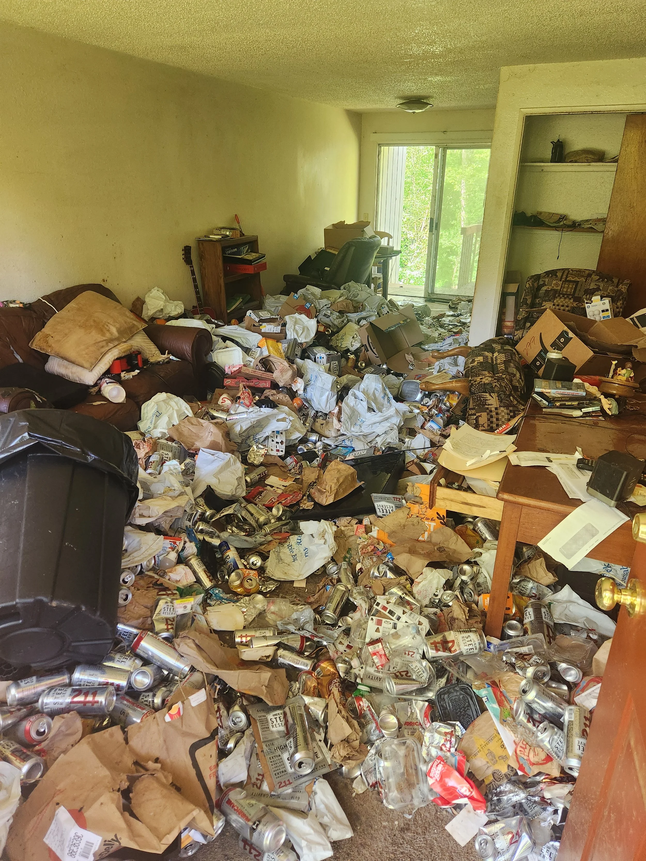 A cluttered living room filled with trash, including crumpled papers, empty cans, and discarded boxes, with a sliding glass door revealing greenery outside.