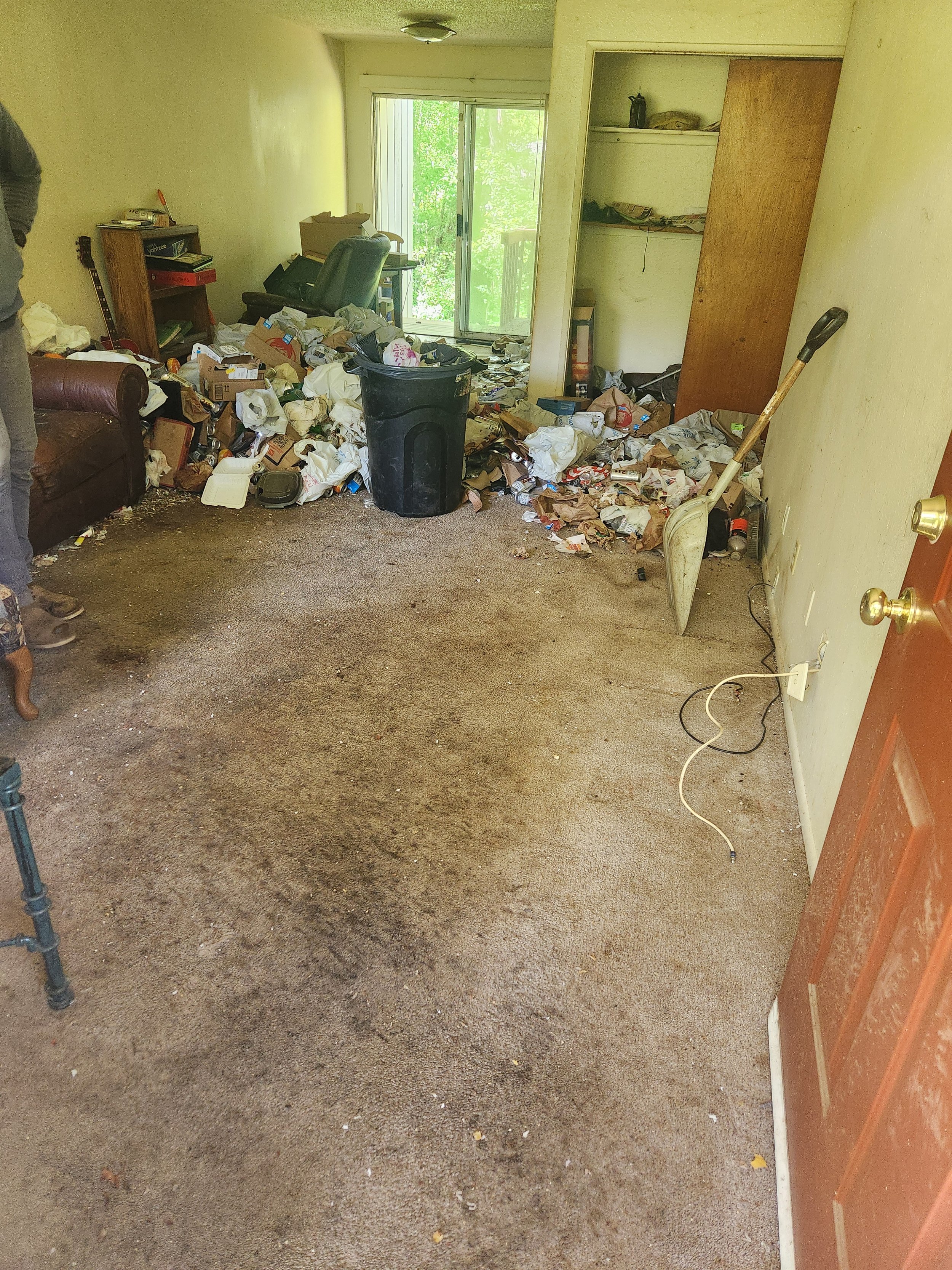 A cluttered living room with a lot of trash and debris scattered on the carpet. There is a sliding glass door leading outside, and various furniture pieces and objects, including a trash can and a shovel, are visible.