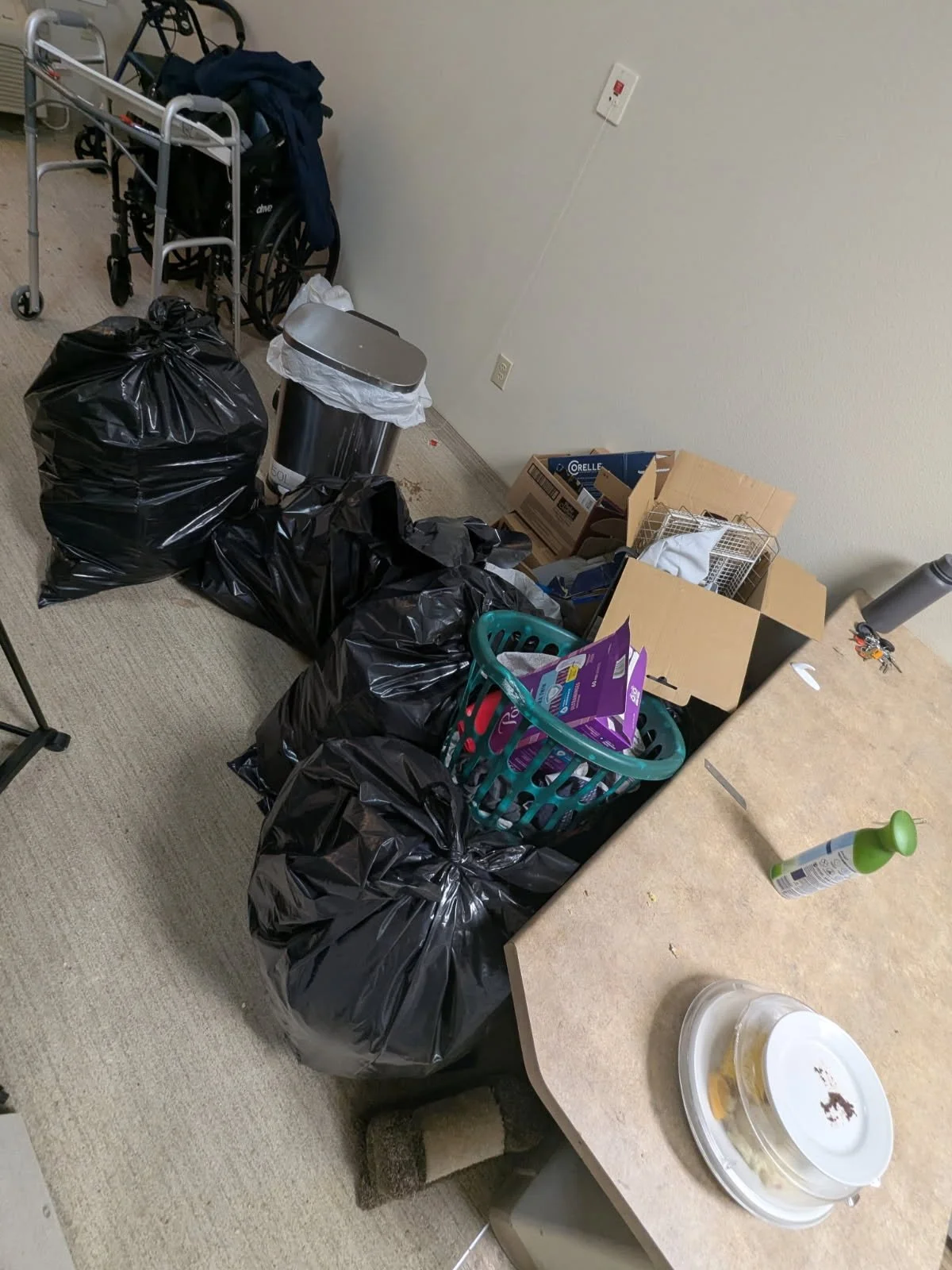 Cluttered room with black trash bags, a cardboard box, a laundry basket filled with clothes or items, a metal trash can, a walker, a countertop with cleaning spray, and scattered small objects.