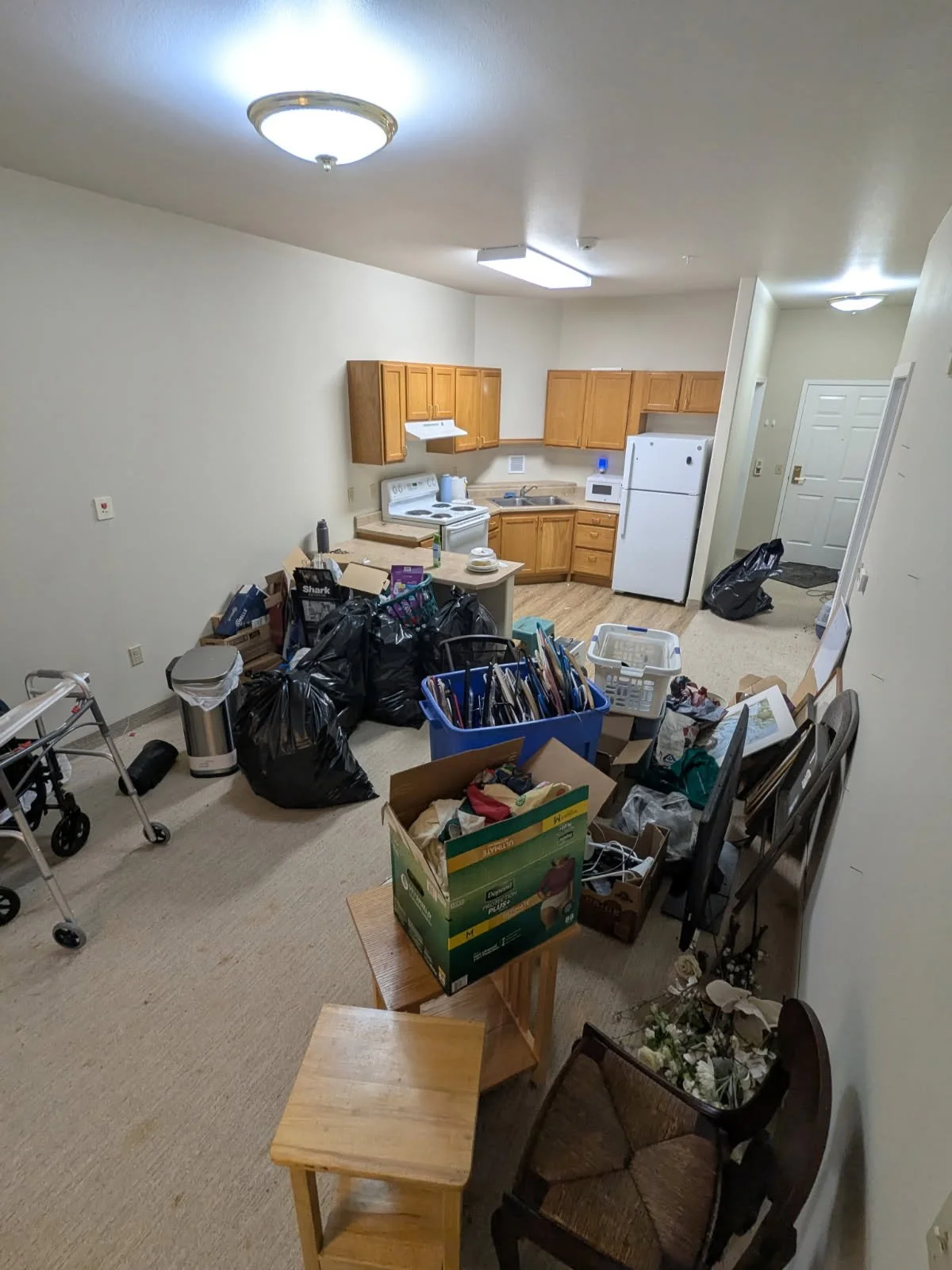 Living room and kitchen area cluttered with black garbage bags, boxes, and miscellaneous items. The kitchen has wooden cabinets, a white stove, microwave, and refrigerator. The walls are plain, and the floor is carpeted.
