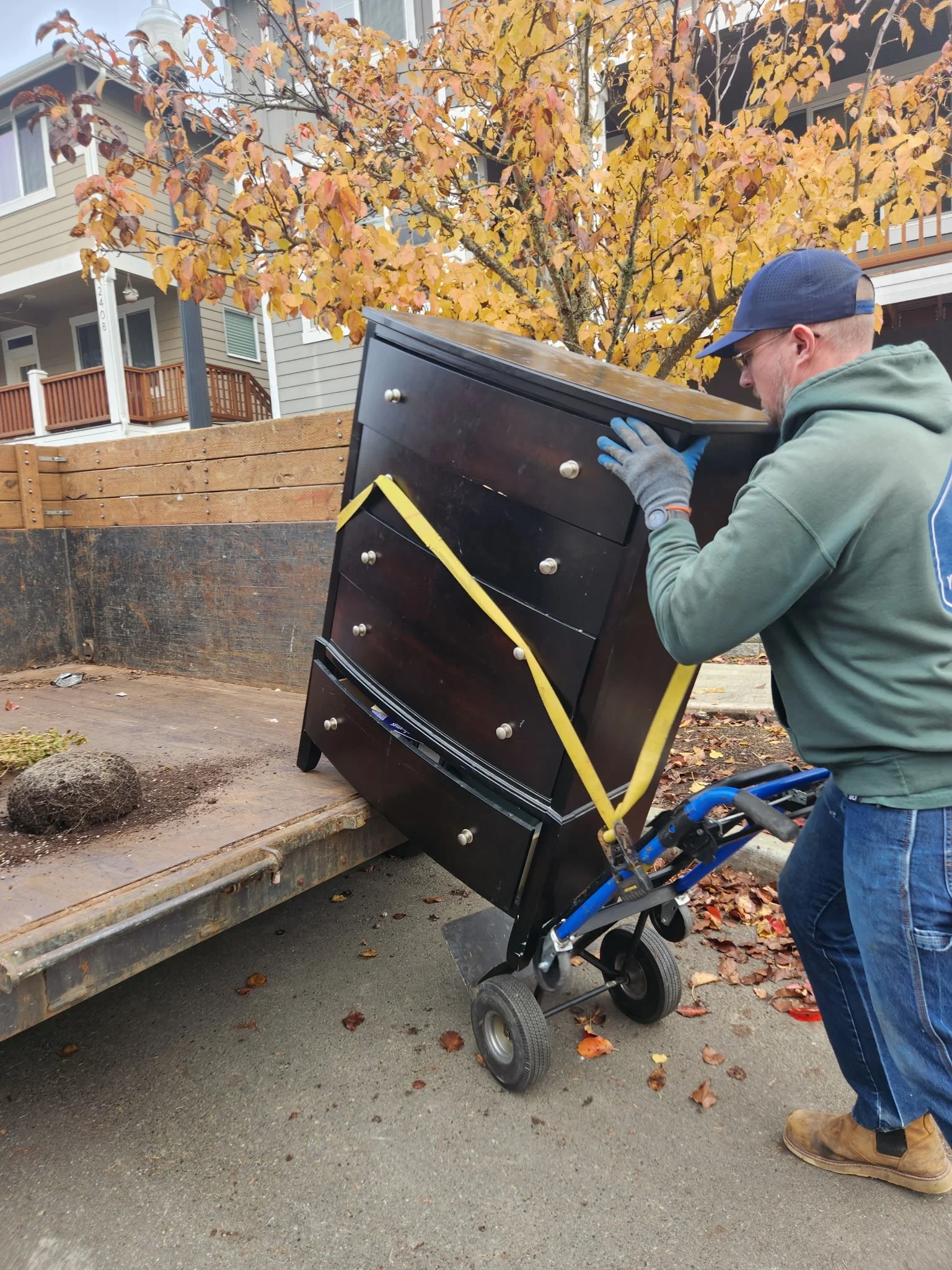 A man with a blue cap, glasses, green hoodie, and work gloves is lifting a black dresser with silver knobs from a flatbed truck using a hand truck or dolly, with a background of an autumn tree with yellow and orange leaves and a residential building.