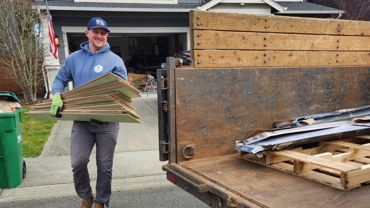 A man wearing a blue hoodie, gray pants, a blue cap, and green gloves is lifting a stack of roofing shingles into the back of a rusty flatbed truck, with a house and garage in the background.