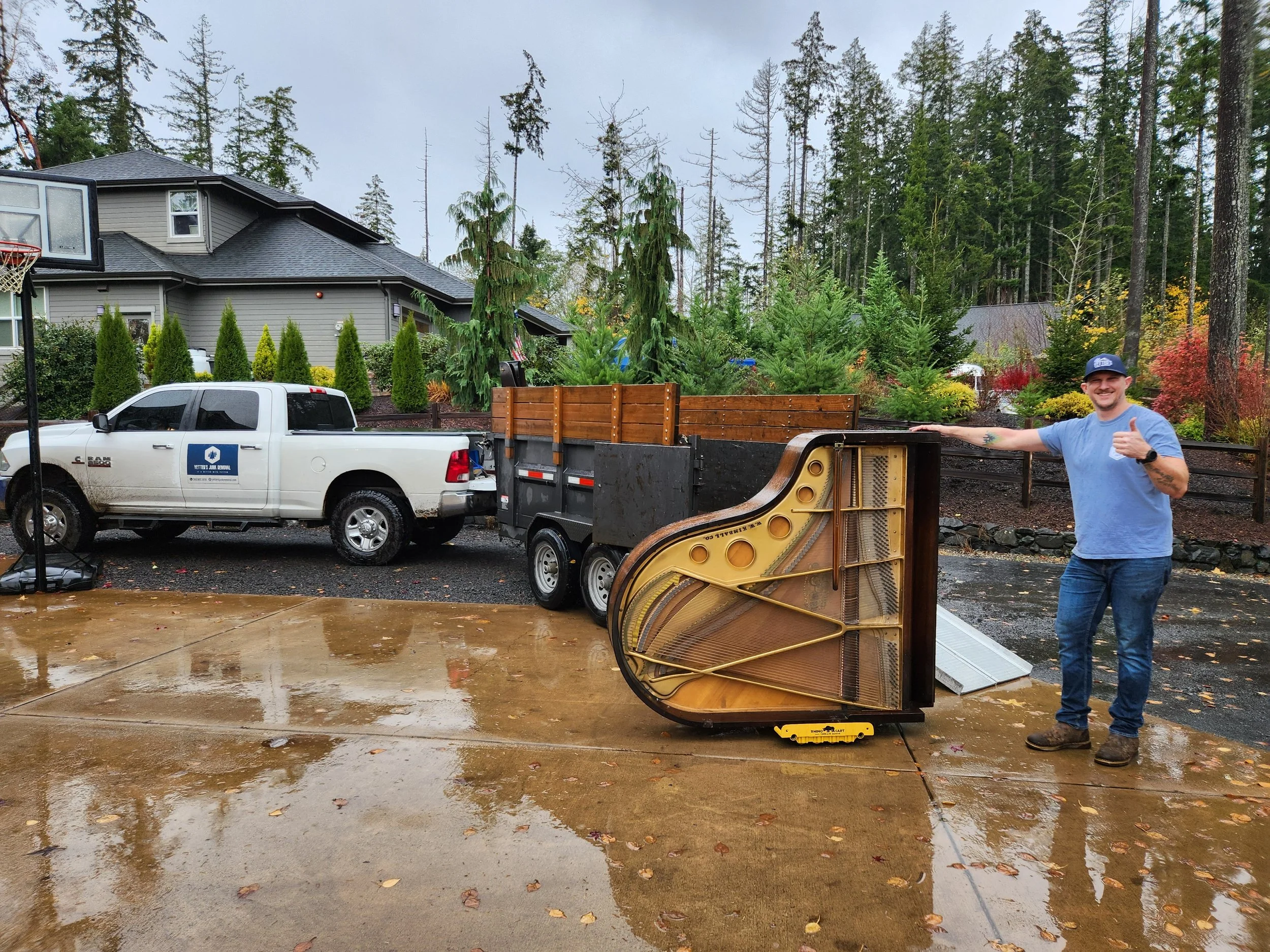 A man smiling and giving a thumbs-up stands next to a large, disassembled piano part on the ground. Behind him, there's a loaded truck hitched to a white pickup truck, with a suburban house, tall trees, and a cloudy sky in the background. The ground is wet with fallen leaves.