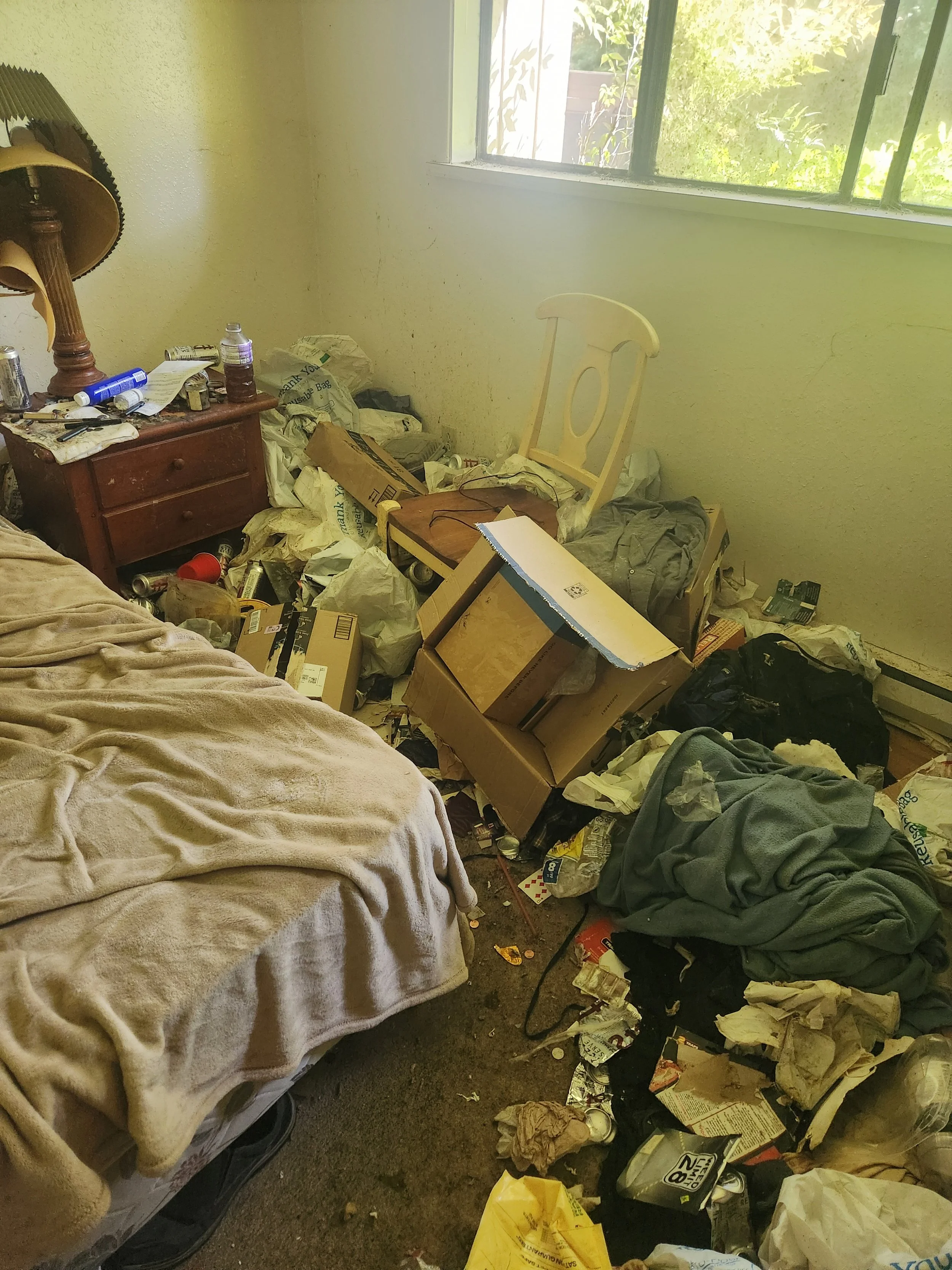 A cluttered bedroom with an unmade bed, trash and clutter on the floor, a small wooden nightstand with a lamp, empty cans, bottles, and papers, and an empty white chair surrounded by trash and cardboard boxes, with a window letting in sunlight.