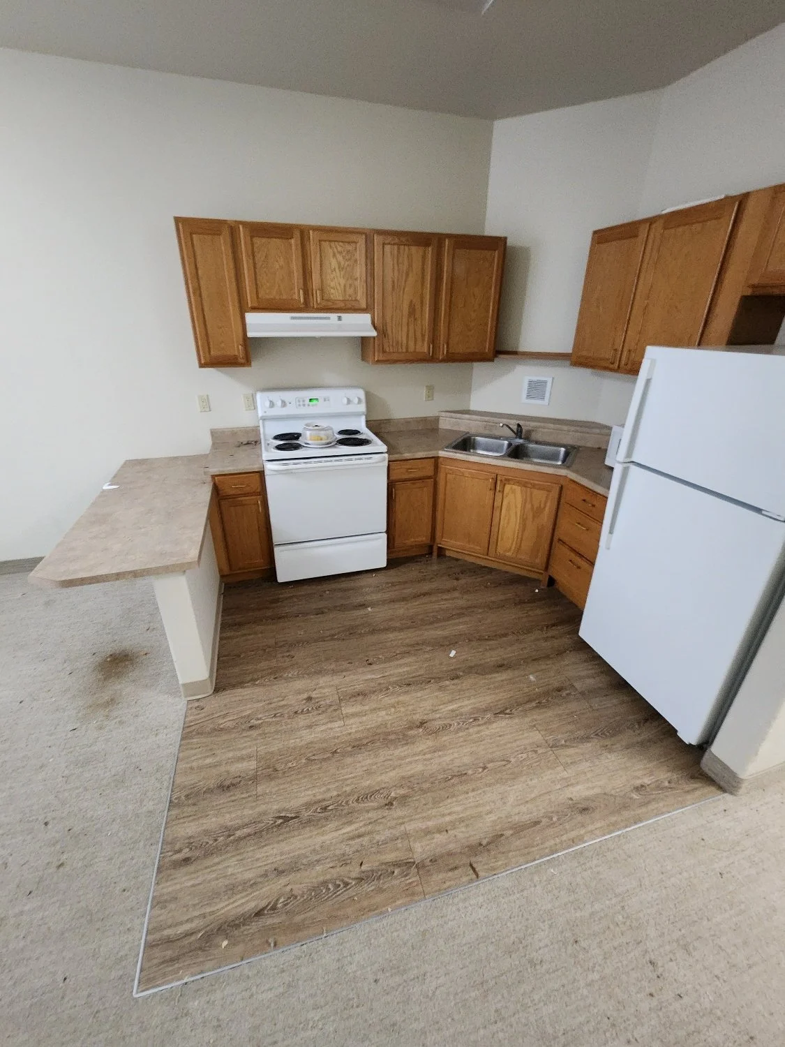 Kitchen with wooden cabinets, white stove, double sink, white refrigerator, and partial view of a countertop extension, with wood and carpet flooring.