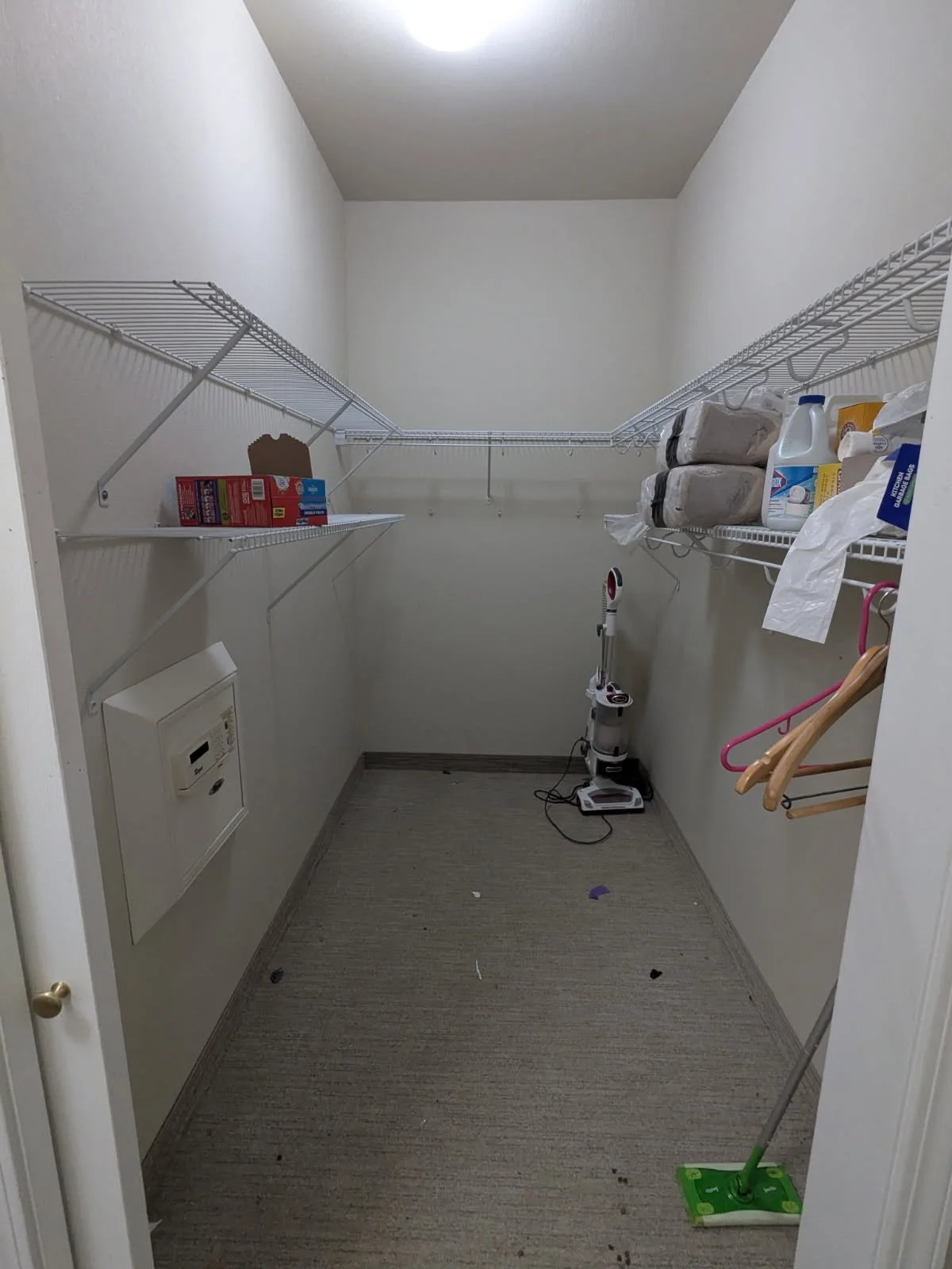 Empty walk-in closet with wire shelves, cleaning supplies, a vacuum cleaner, hangers, and a small safe on the wall. Floor has some dirt and debris.