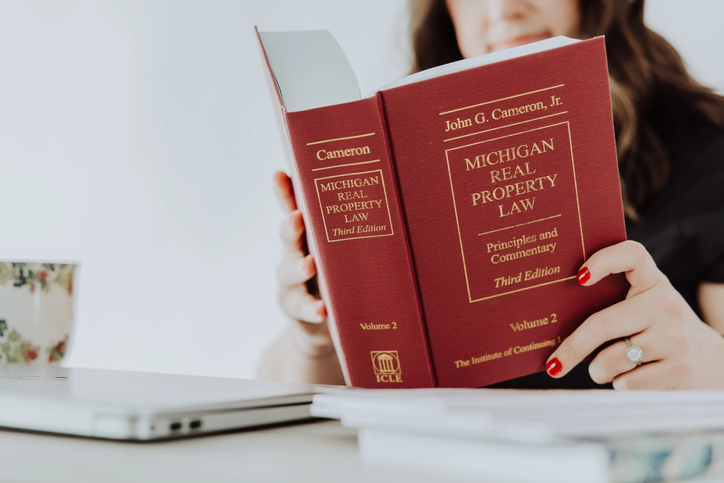 A woman holding a red book titled "Michigan Real Property Law" with her left hand, showing a diamond ring on her finger, while smiling. The book is the third edition, volume 2, and published by ICLE.