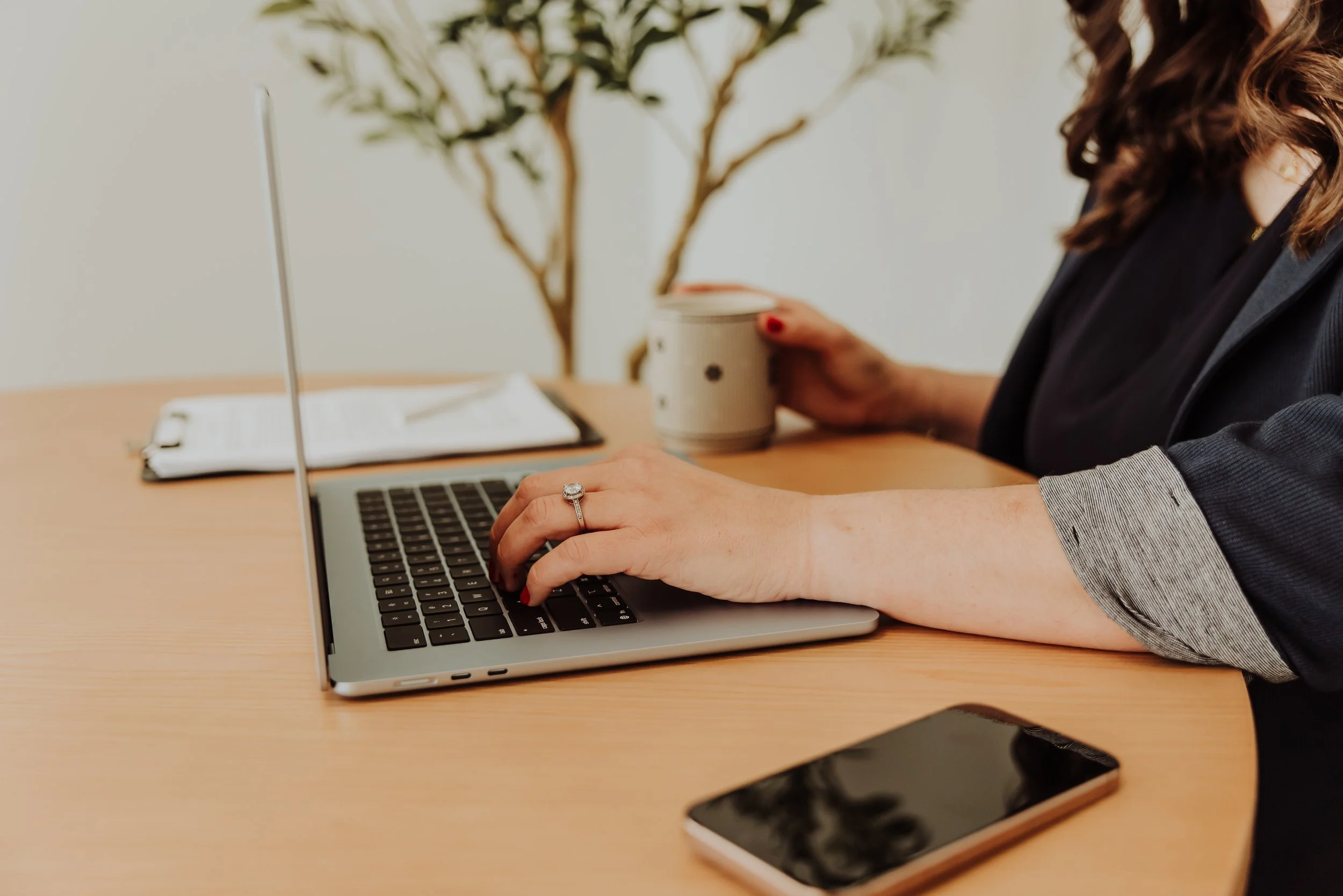 Woman working on a silver laptop at a wooden desk, holding a mug in her left hand. A smartphone is on the desk, and a clipboard with papers is in the background, with a tall plant nearby.