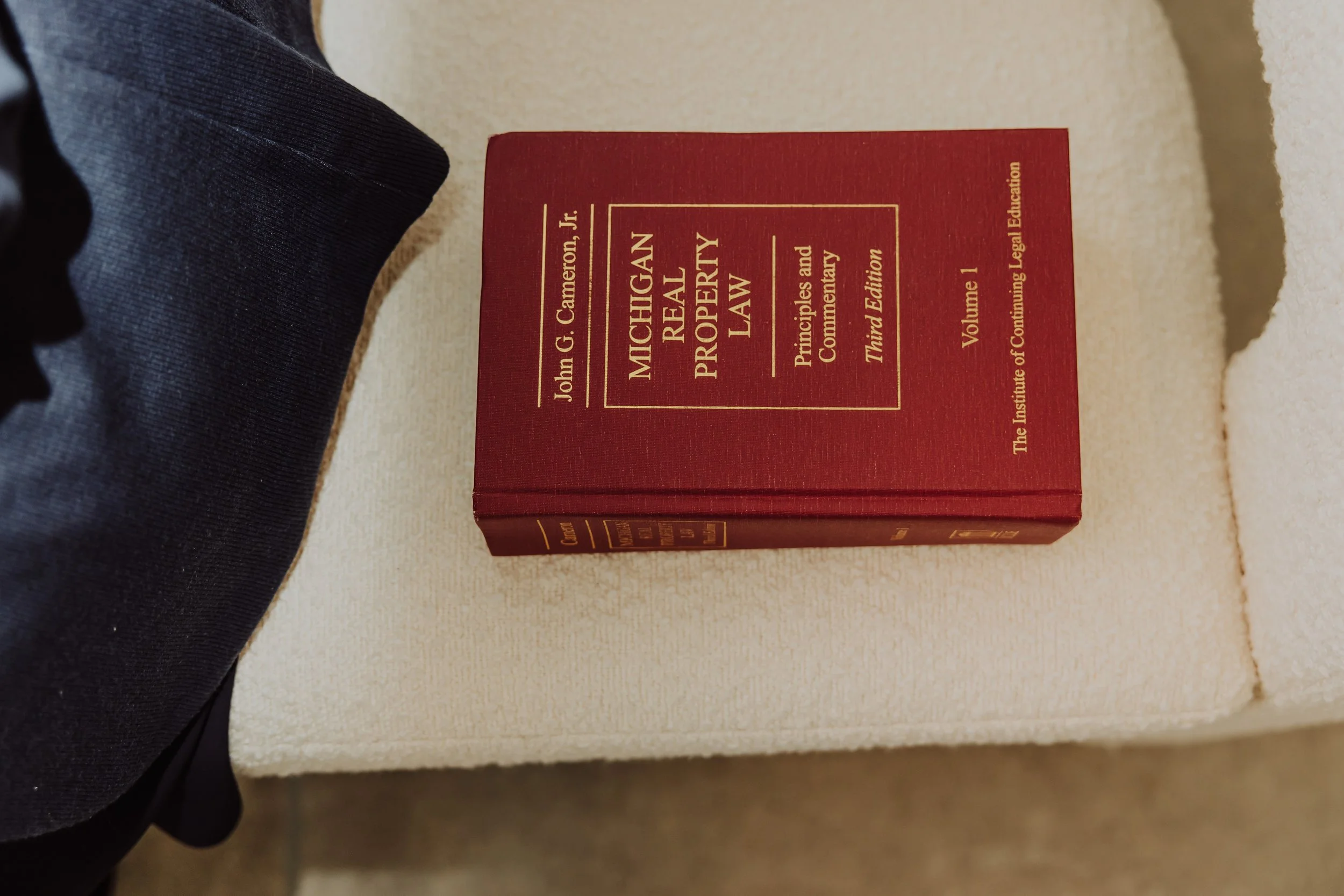 Red hardcover book titled "Michigan Real Property Law" by John G. Cameron Jr., third edition, volume 1, lying on a cream-colored surface, with part of a dark clothing sleeve visible on the left.